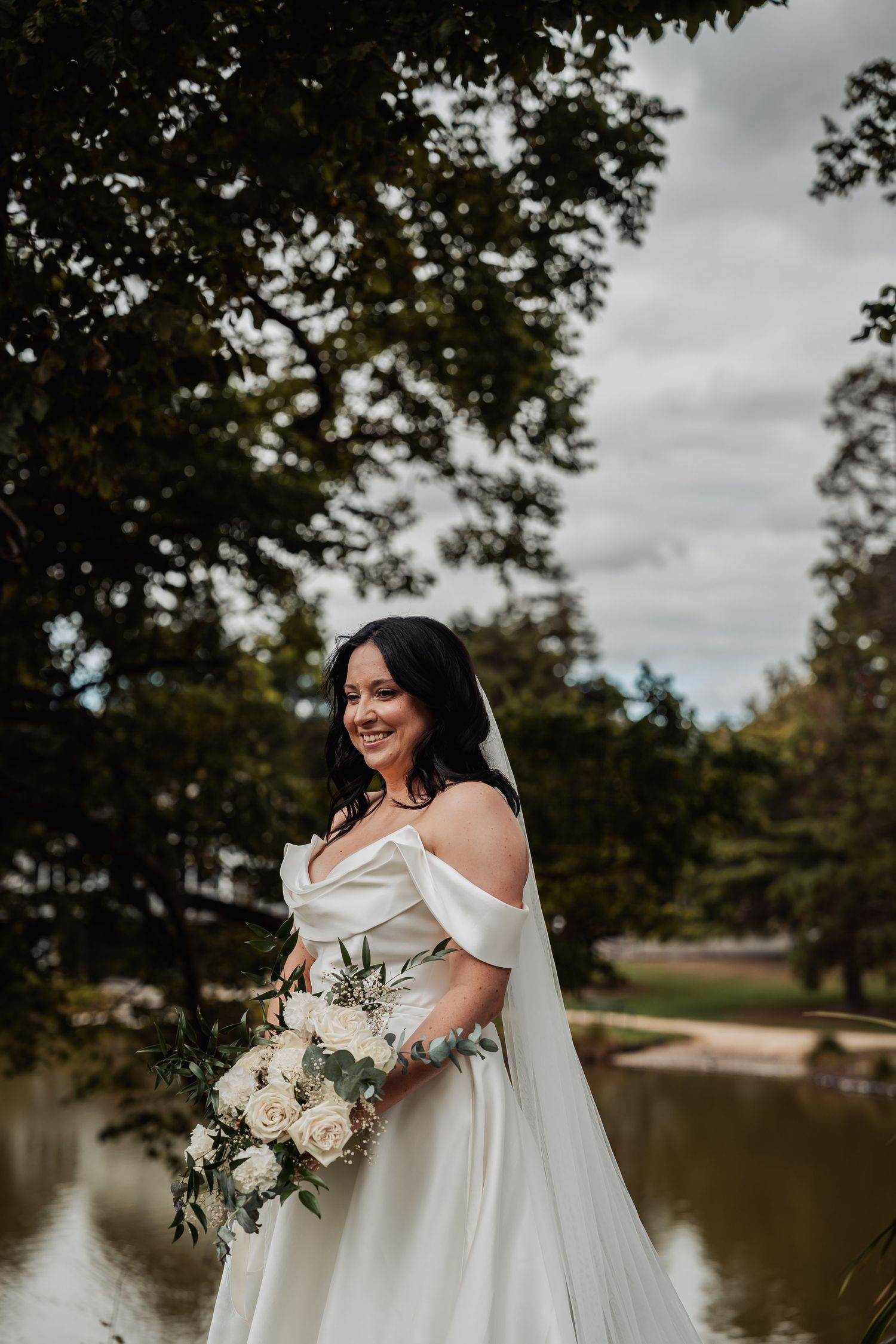 A smiling bride in an off-shoulder white gown holds a white rose bouquet beside a tranquil pond.