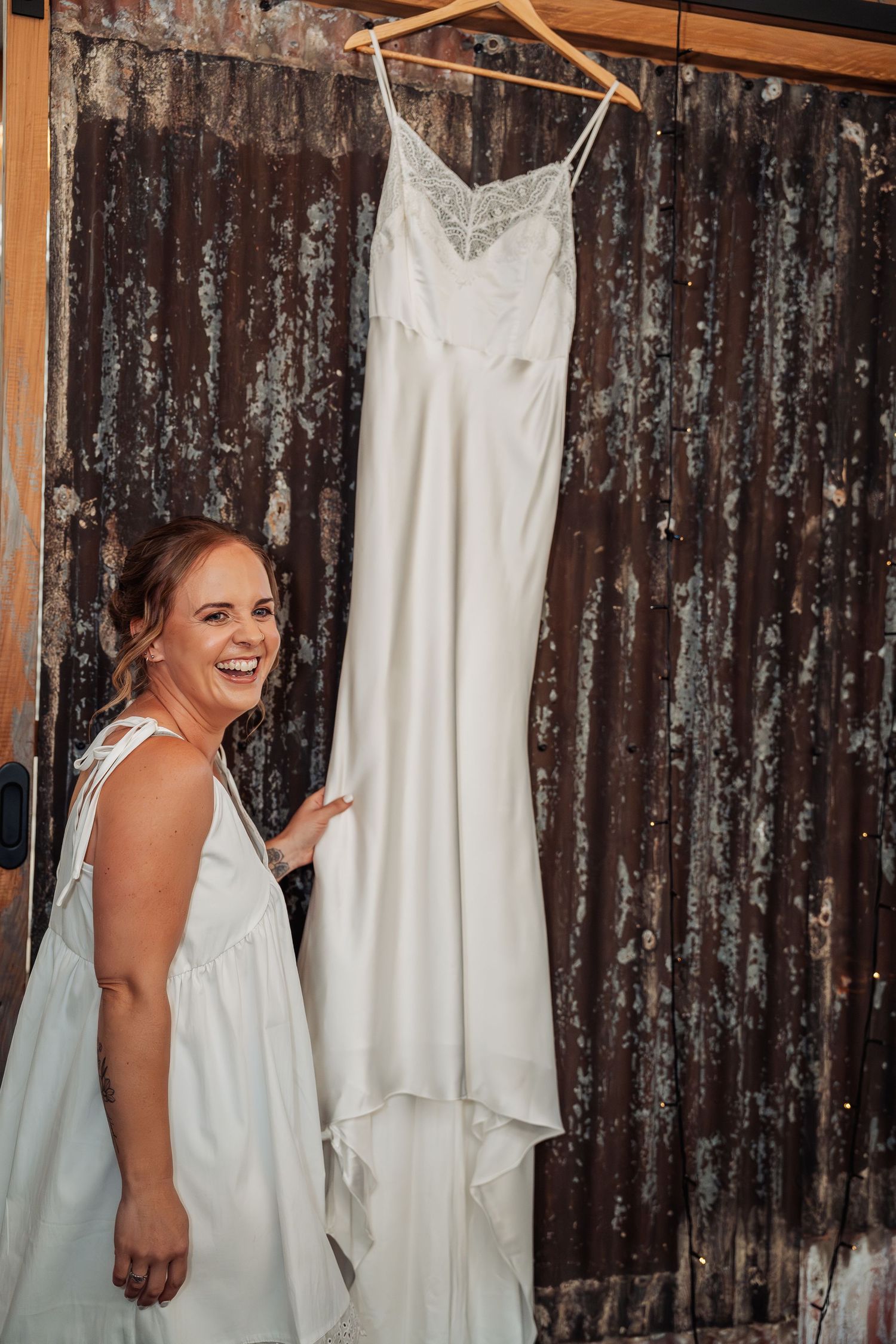 Bride laughing beside her elegant white satin wedding dress hanging on a rustic wooden door.