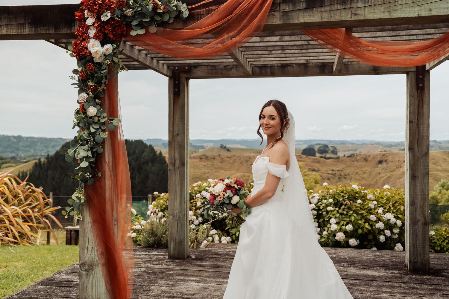 Bride in off-shoulder white gown holding bouquet at outdoor ceremony with scenic landscape.
