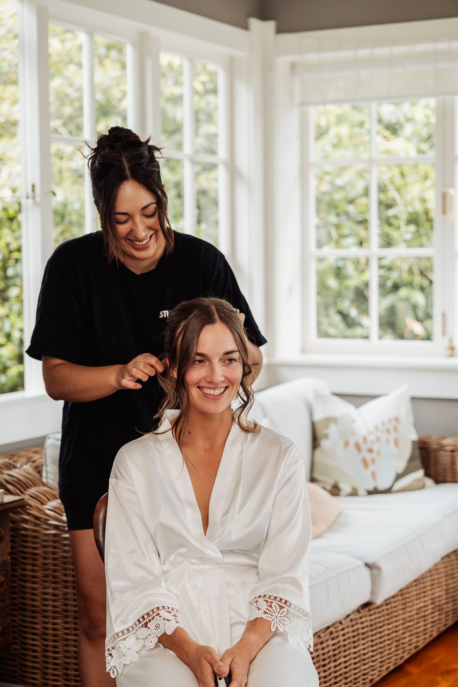 Smiling bride in white robe gets hair styled by hairdresser in bright room with large windows.