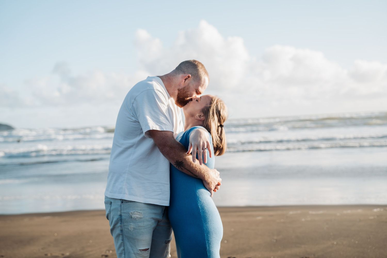 Couple sharing a romantic embrace on sandy beach at sunset with ocean waves in background.