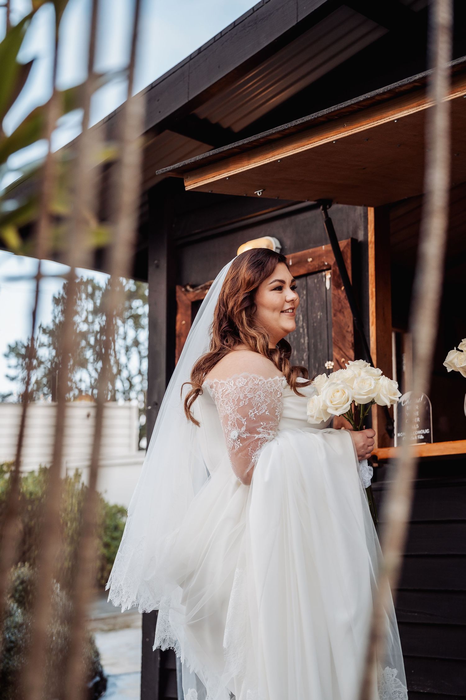 A bride in a lace-sleeved white gown holding a bouquet stands under a wooden structure outdoors.