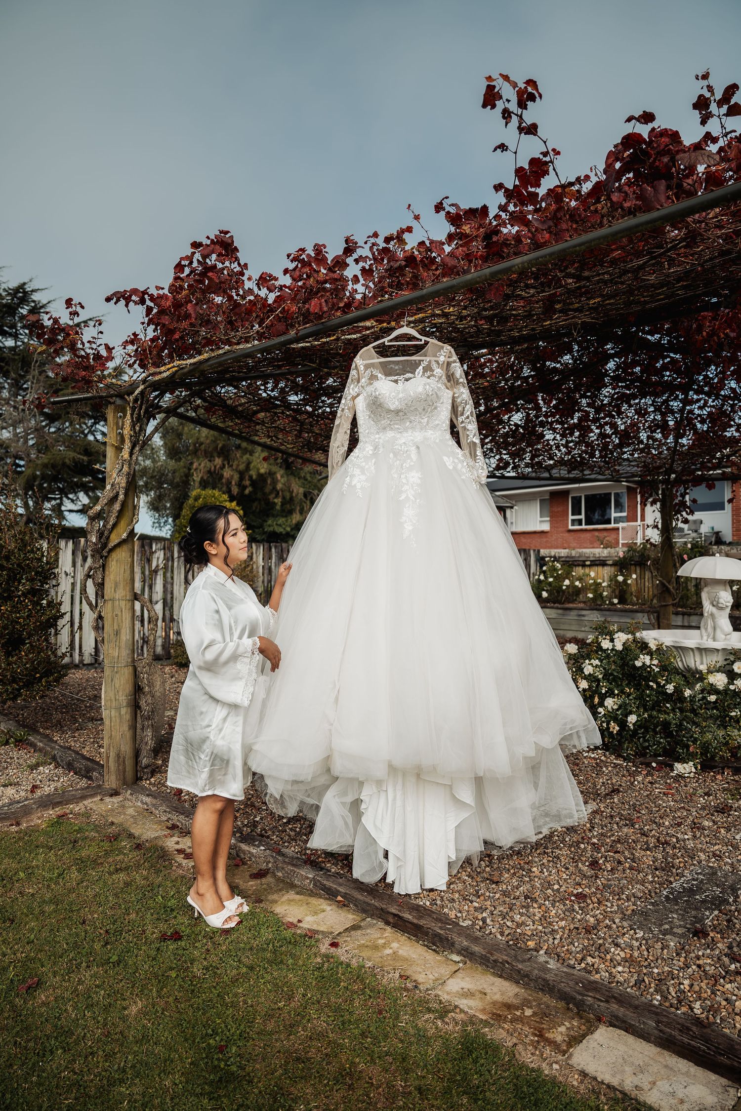 Bride in white robe admiring her lace ball gown wedding dress hanging outdoors under a pergola.