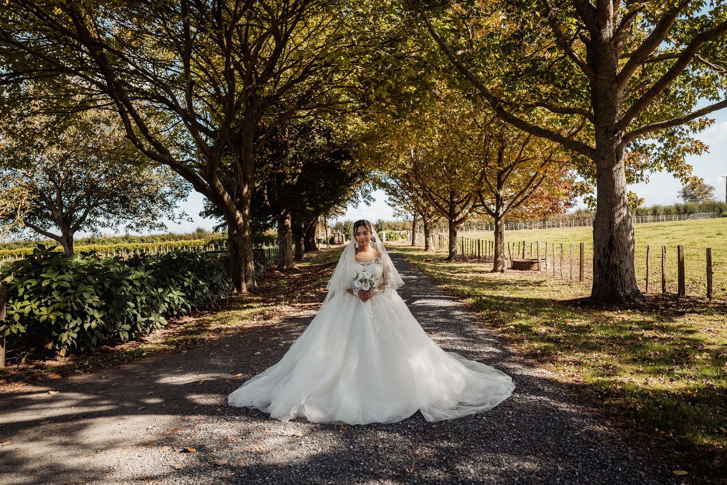 Bride in full white gown and veil stands on scenic tree-lined path holding white bouquet.