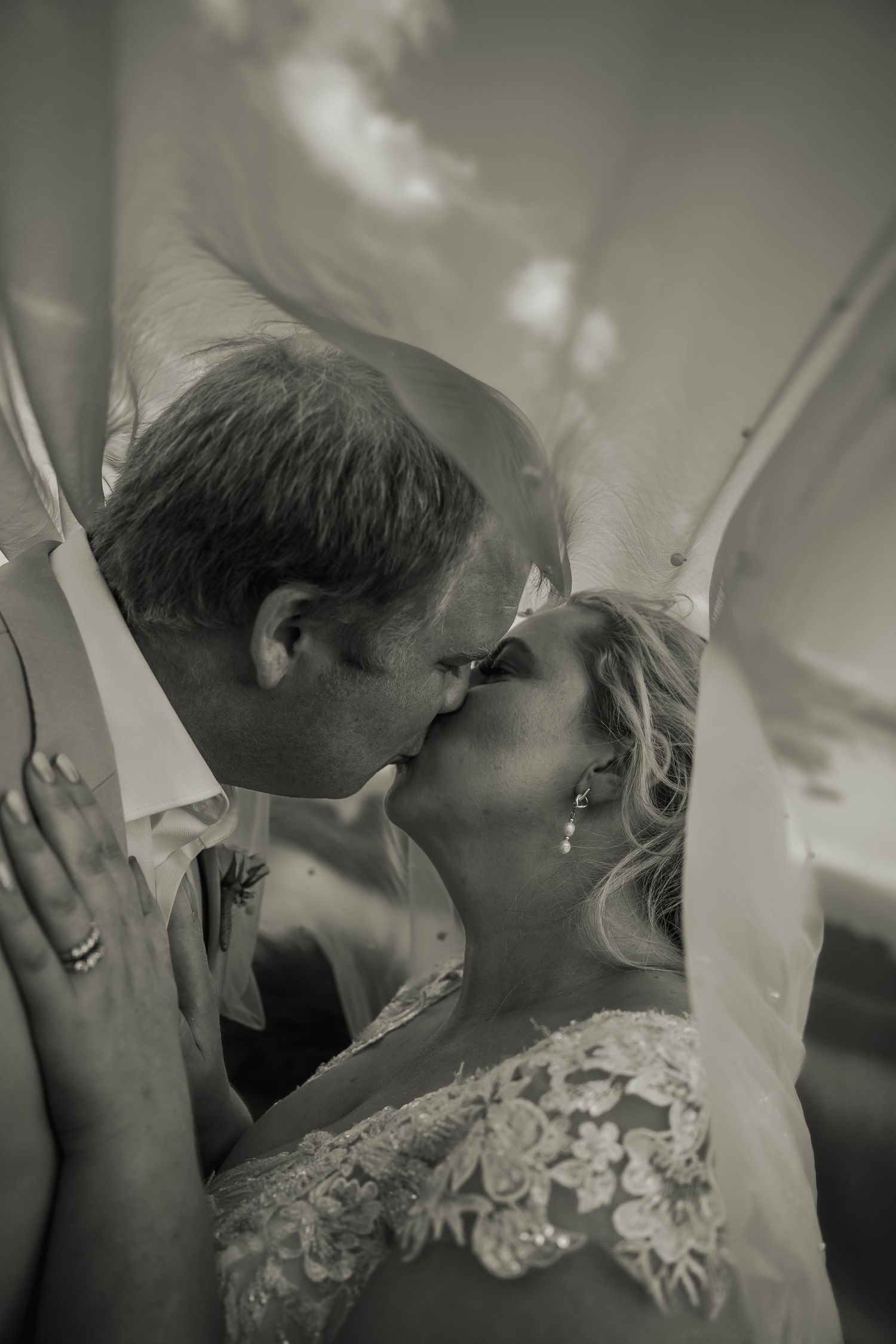 Bride and groom share a romantic kiss under a flowing veil in a dreamy black and white wedding portrait.