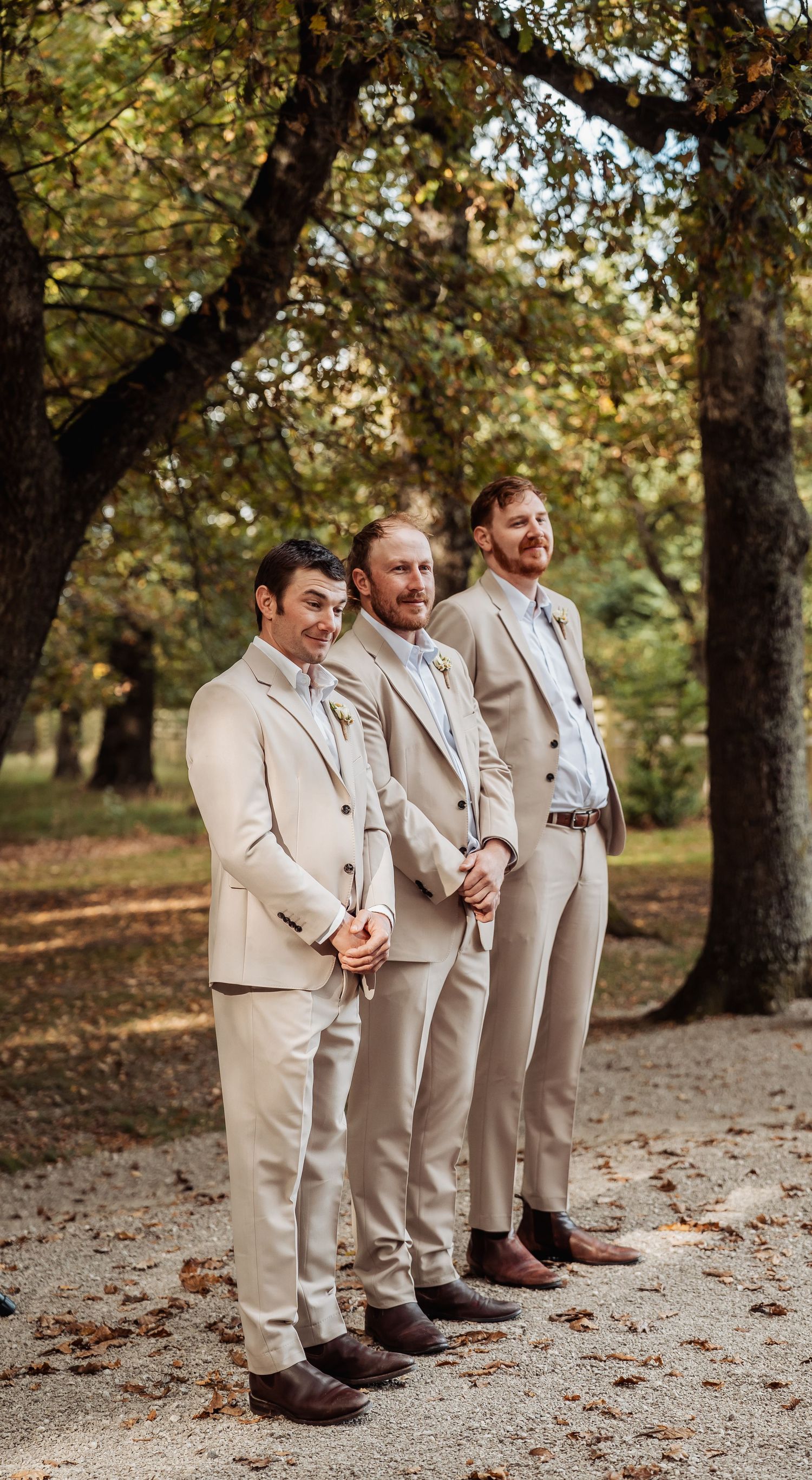 Three groomsmen in matching tan suits standing together under autumn trees outdoors.
