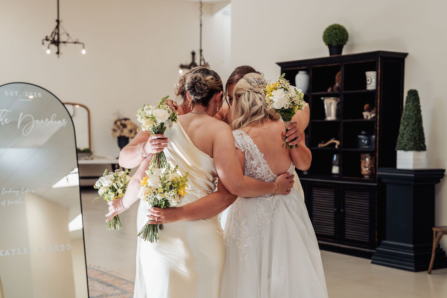 Two brides in white gowns embrace while holding yellow and white bouquets in front of a mirror at their wedding venue.