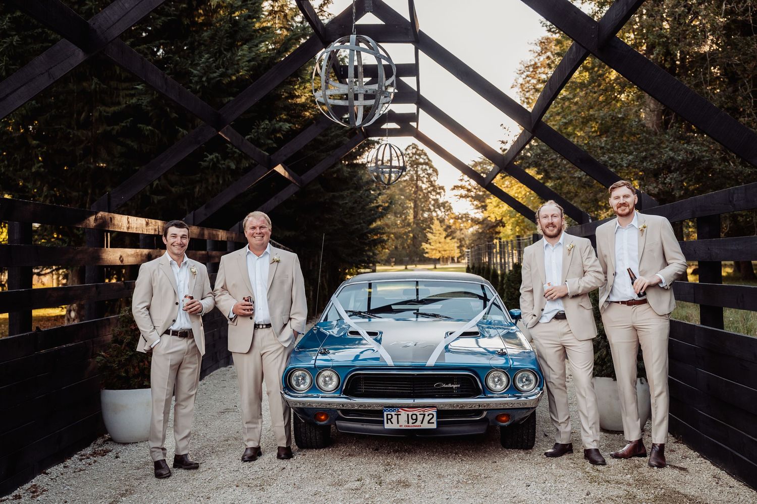Four groomsmen in tan suits pose with a classic blue muscle car at a rustic wedding venue.