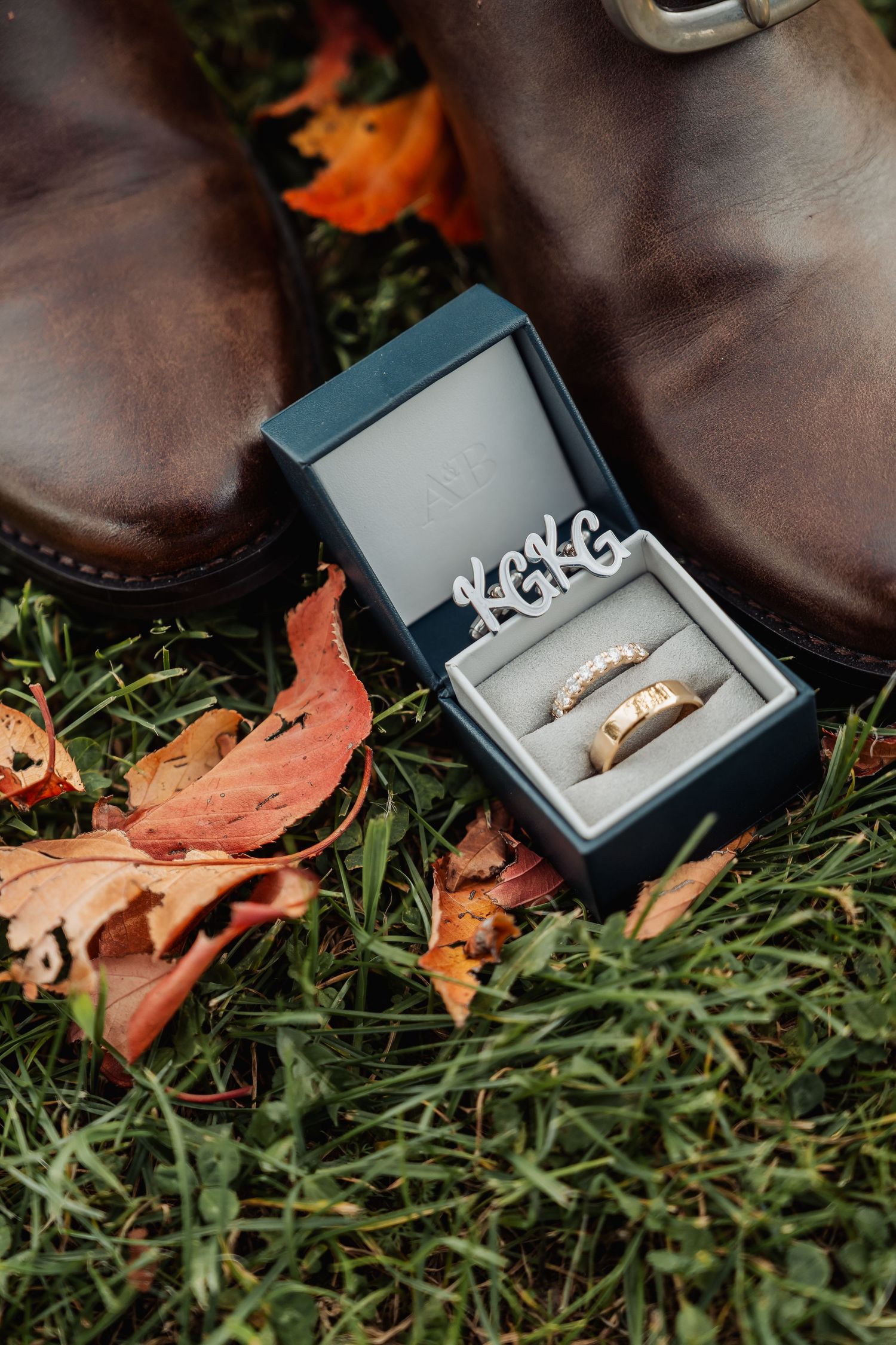 Close-up of wedding rings in a box beside brown leather boots on fall leaves.