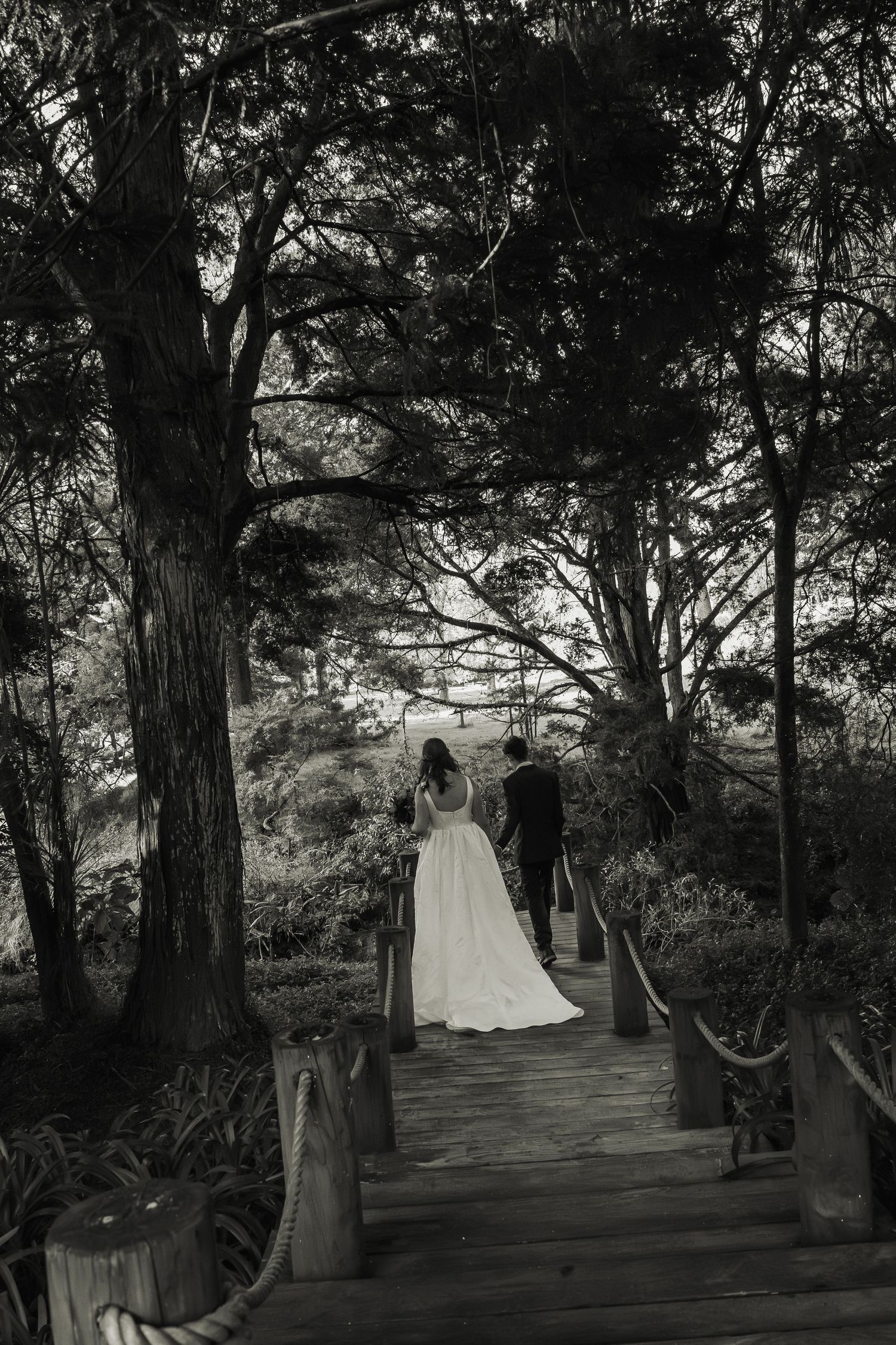 Bride and groom descend wooden stairway through dense forest in black and white photo.