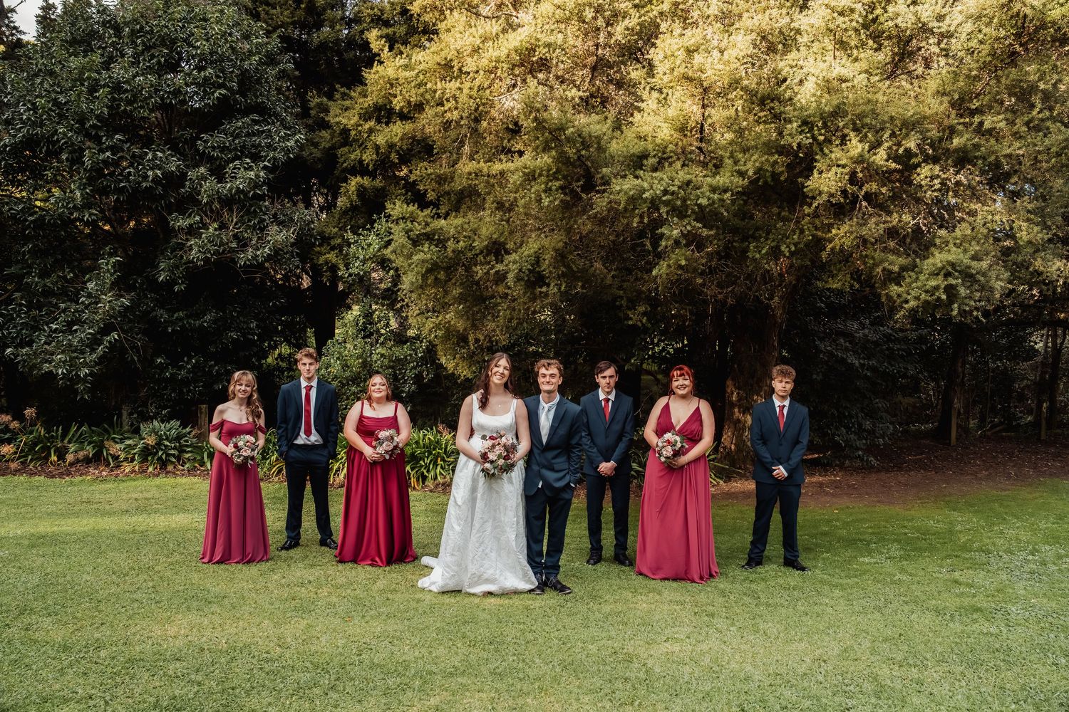 Wedding party poses on green lawn, bride in white gown flanked by bridesmaids in burgundy and groomsmen in navy suits.
