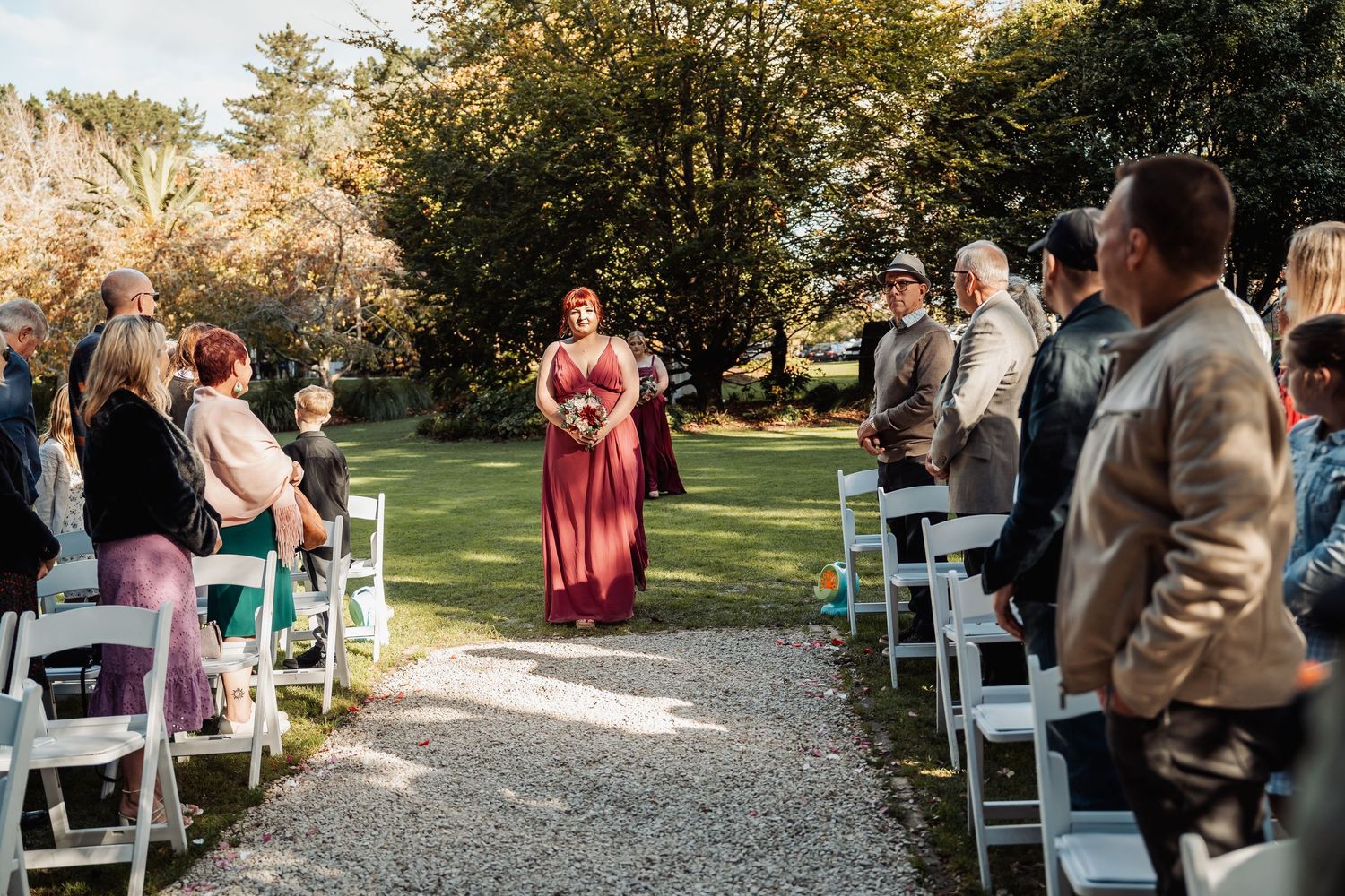 Bridesmaid in burgundy gown walks down outdoor wedding aisle lined with white chairs and seated guests.