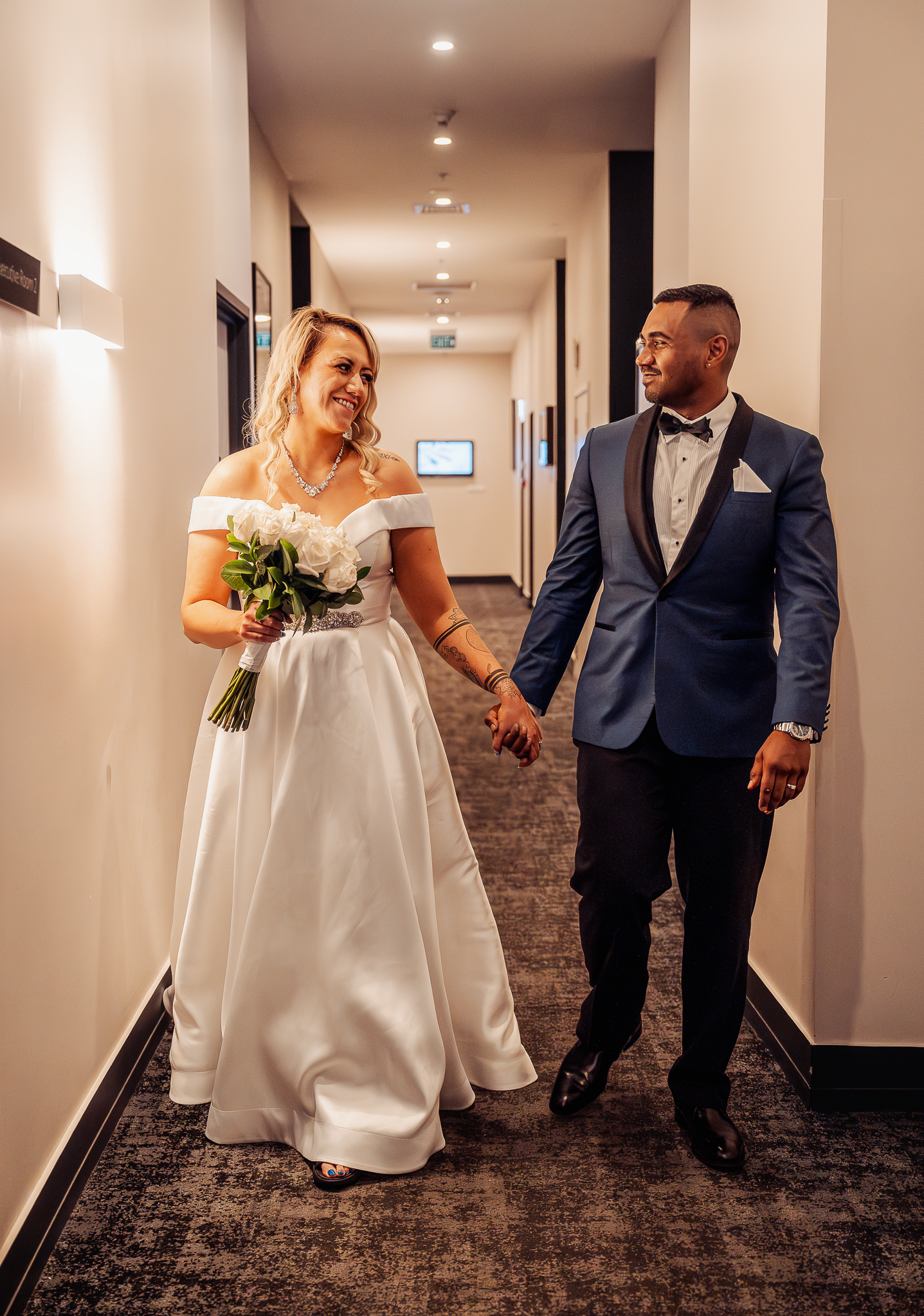 Couple walks hand in hand down hotel hallway in elegant wedding attire.