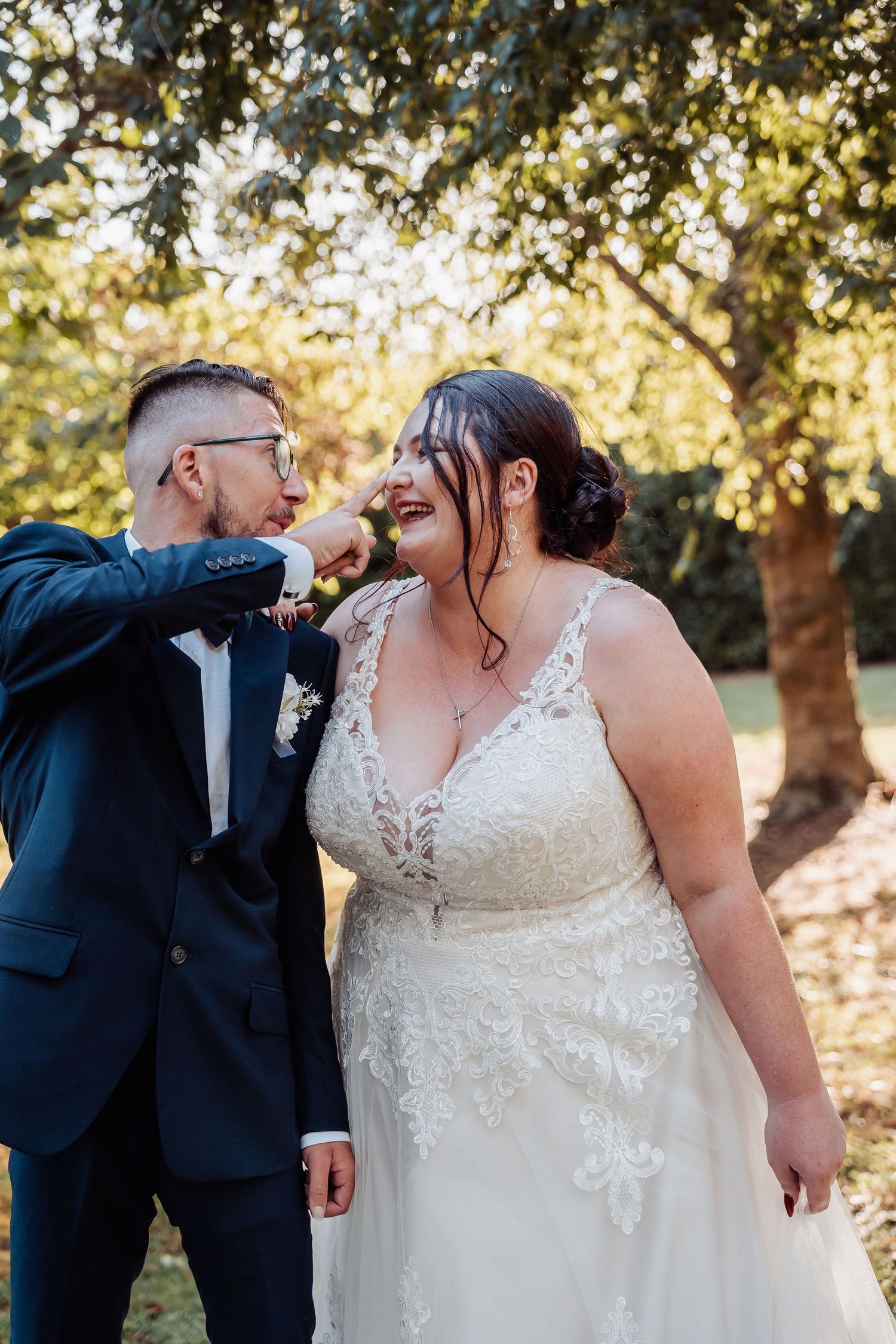 Joyful wedding couple sharing a lighthearted moment in front of trees with golden sunlight.