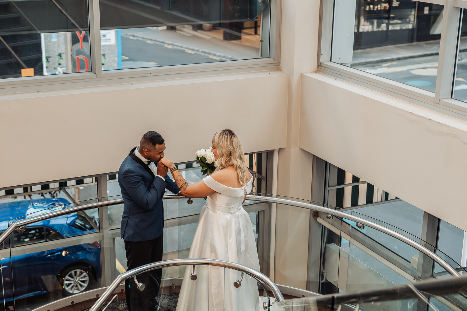 Couple in wedding attire shares a romantic moment on a modern glass-railed staircase.