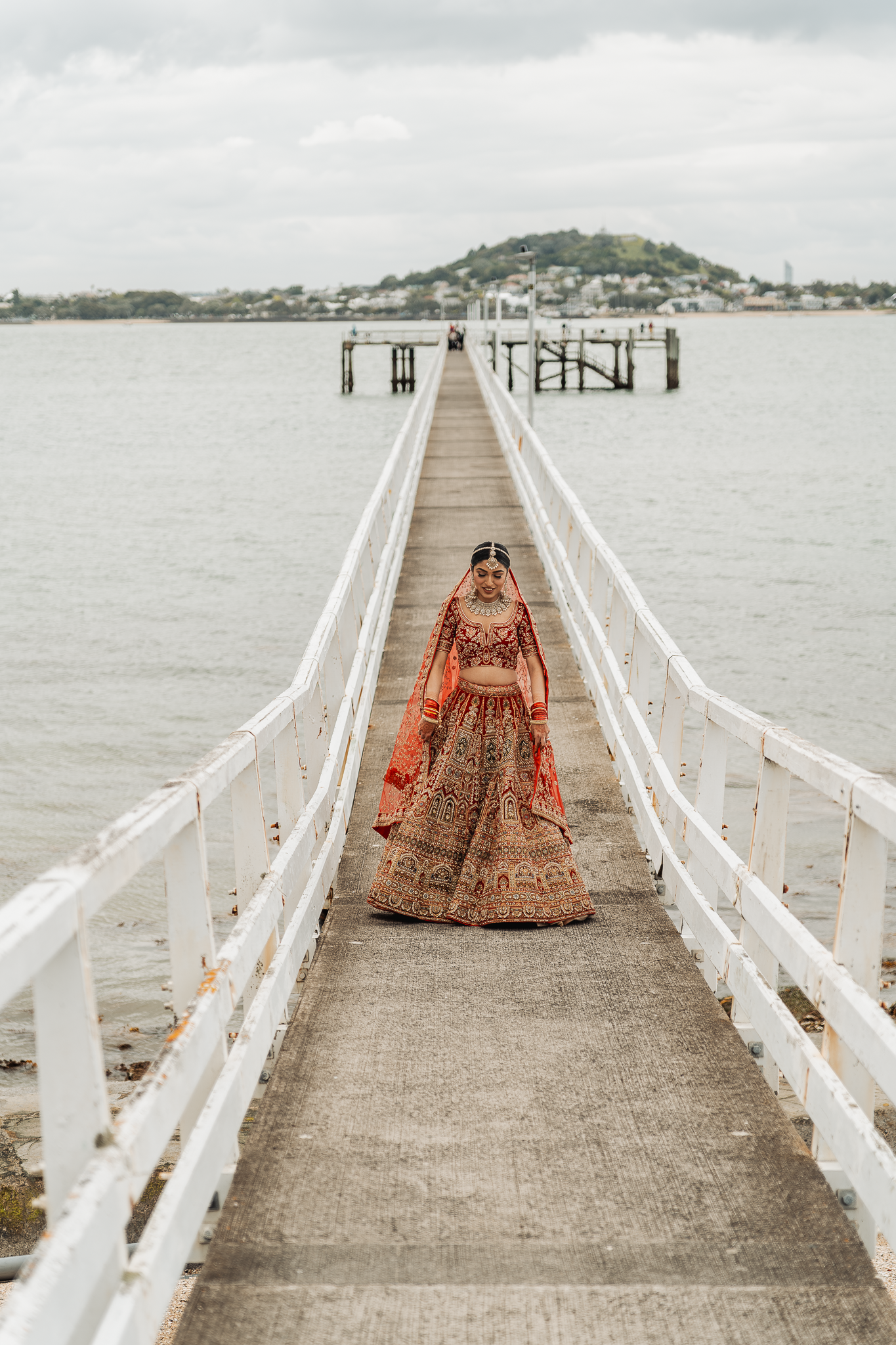 Bride in traditional Indian wedding attire stands on a long white pier over water.