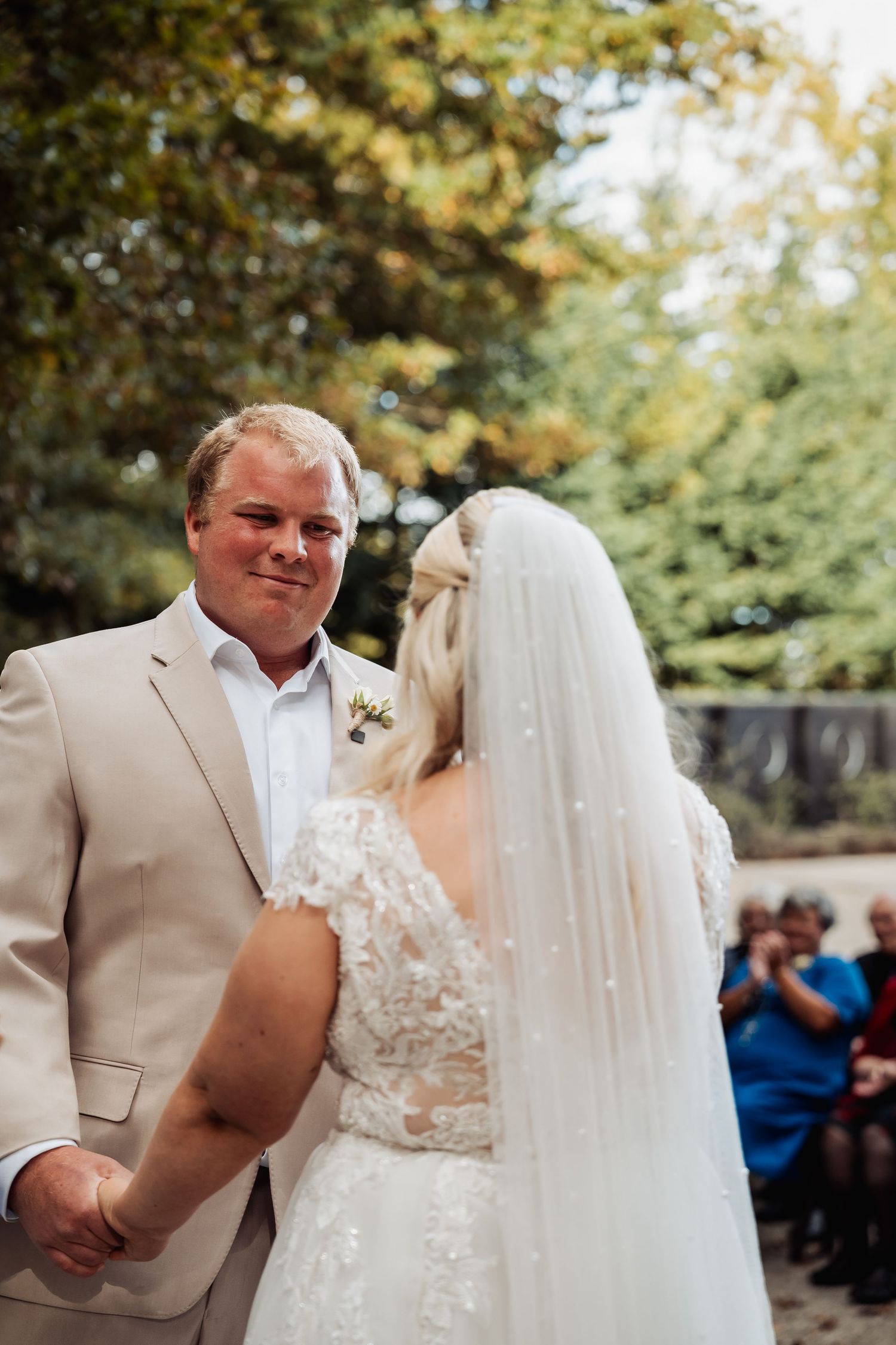 Groom in tan suit smiles at bride in lace wedding dress and veil during outdoor wedding ceremony.