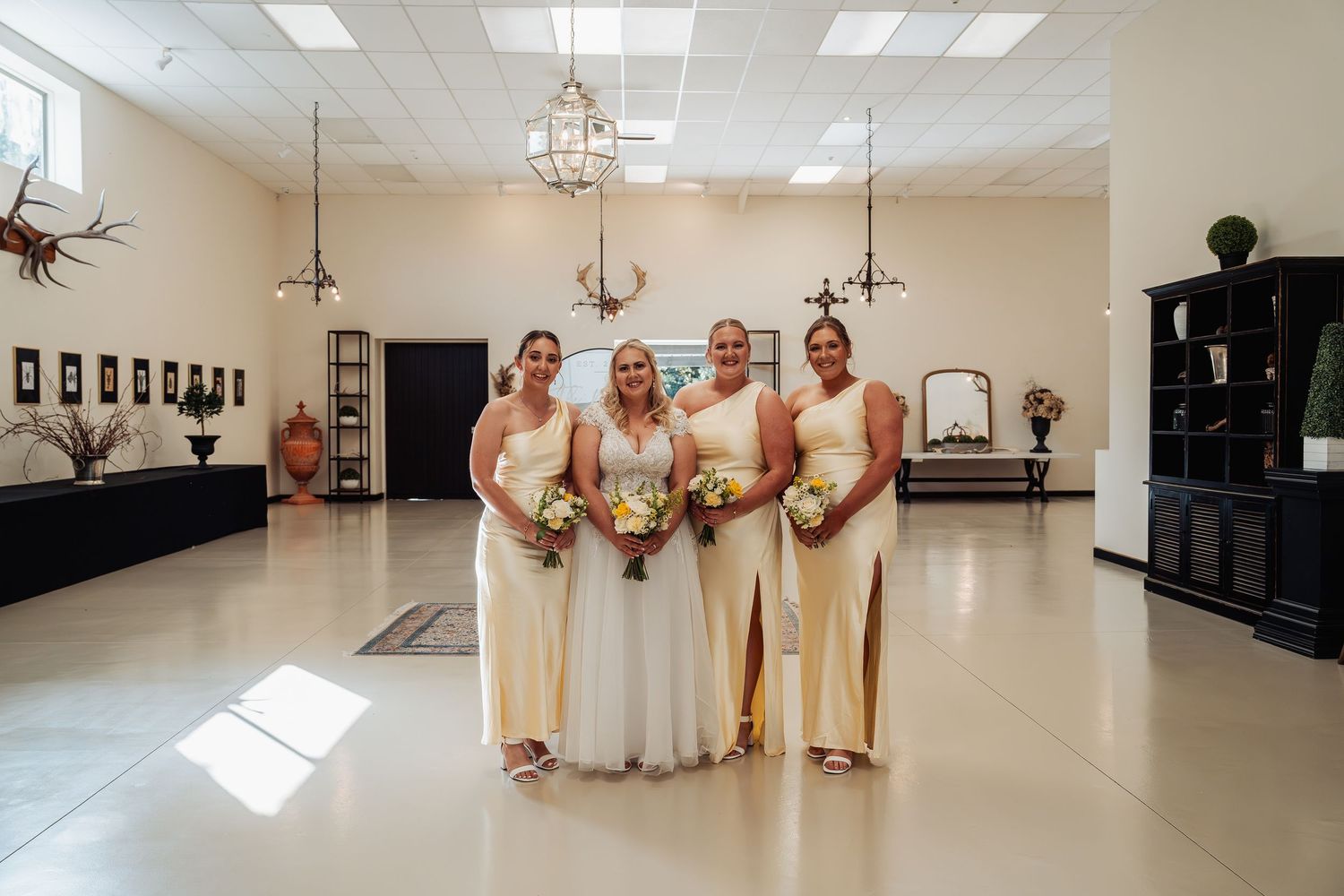 Bride and three bridesmaids in yellow gowns pose with bouquets in an elegant white wedding venue hall.