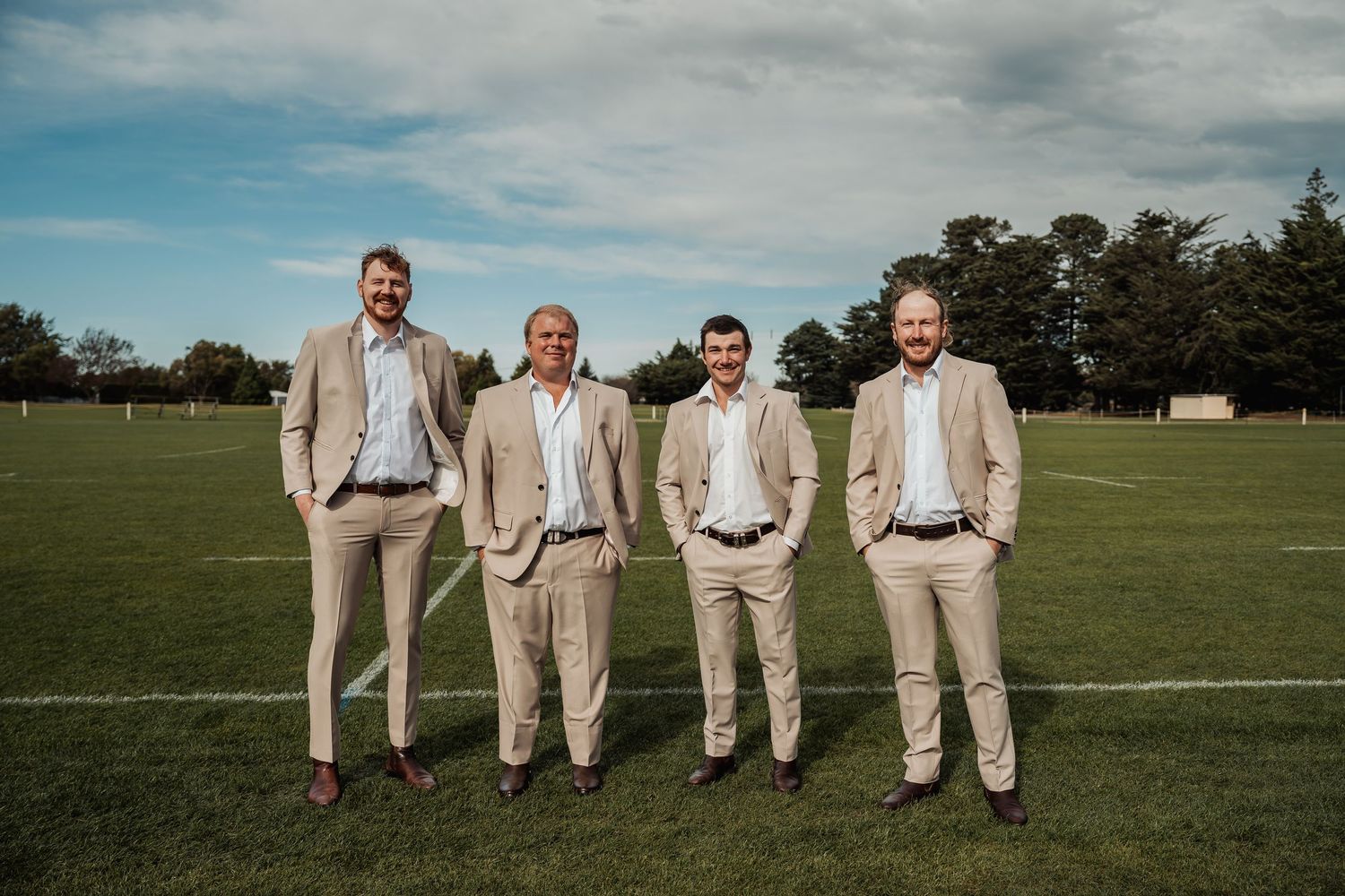 Four men in matching beige suits smiling together on a green sports field under a partly cloudy sky.
