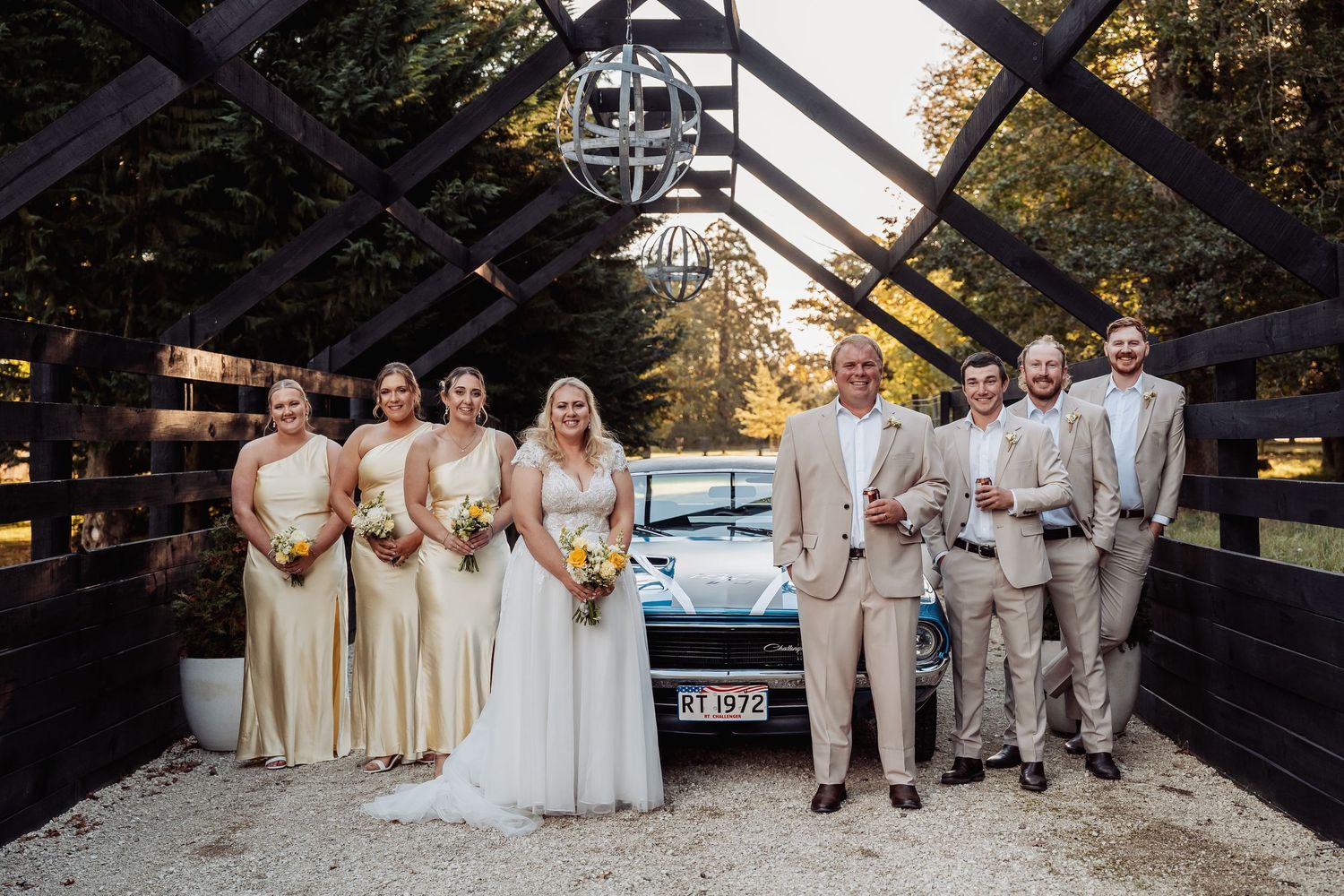 Wedding party posing under wooden arch with vintage car at rustic outdoor venue.