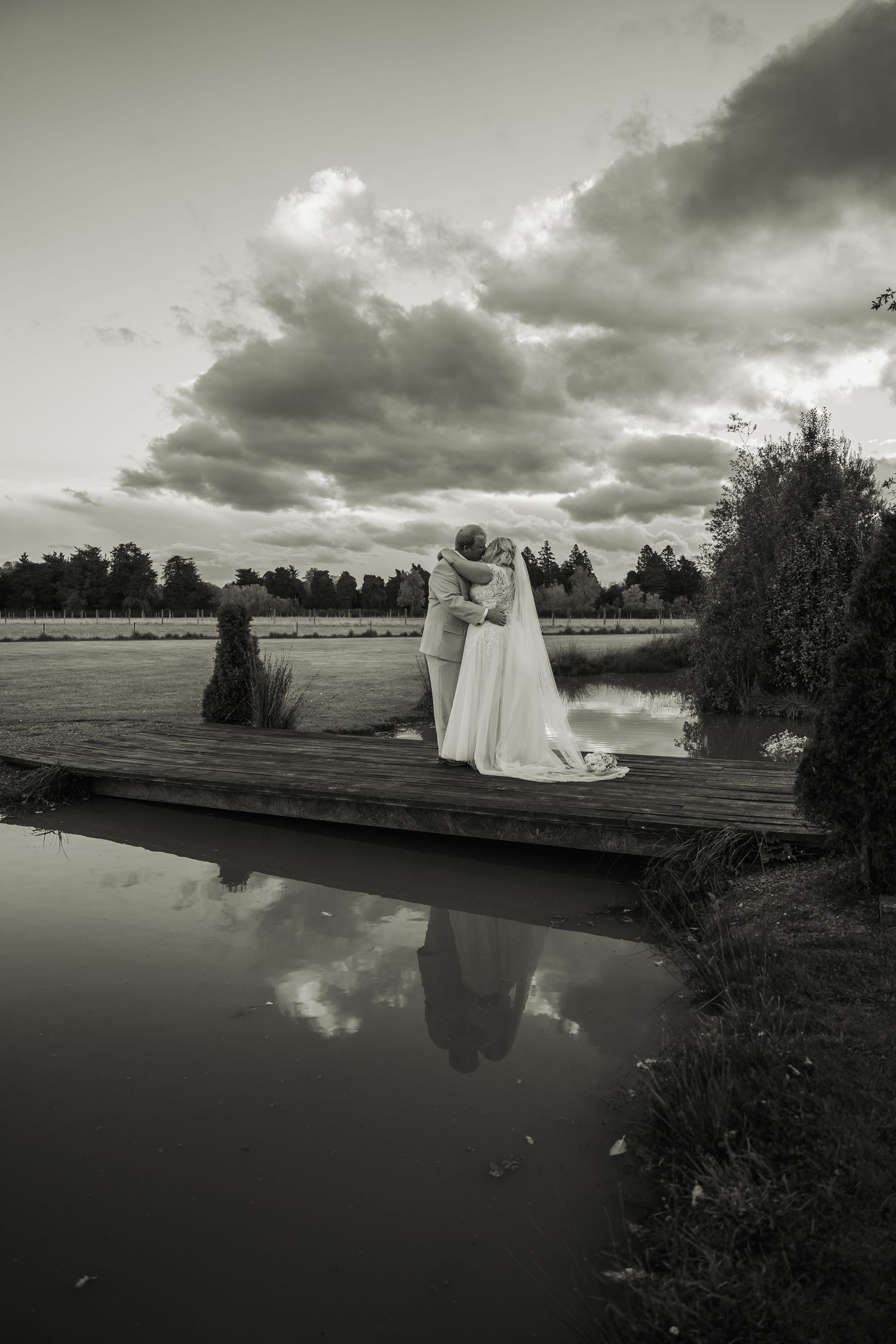 Black and white photo of bride and groom kissing on wooden dock by pond.