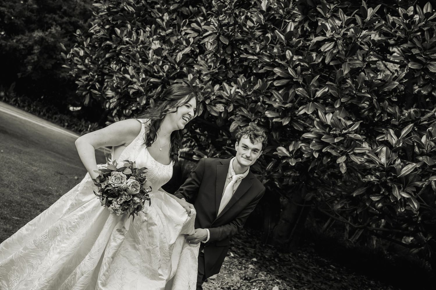 Black and white photo of bride holding bouquet and groom walking beside lush garden hedge.