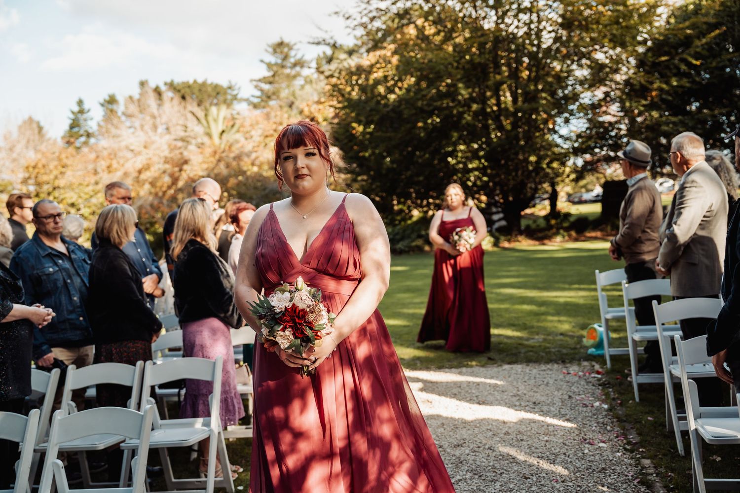 Bridesmaid in burgundy gown walking down outdoor wedding ceremony aisle with bouquet.