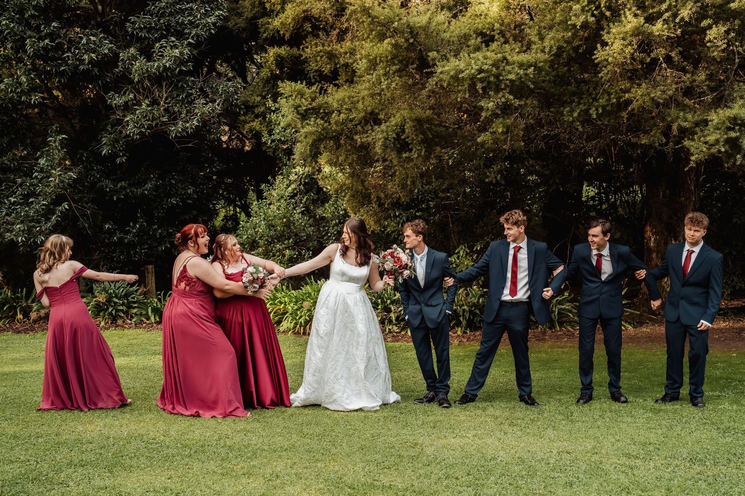 Playful wedding party laughing and pulling each other on a green lawn surrounded by lush trees.
