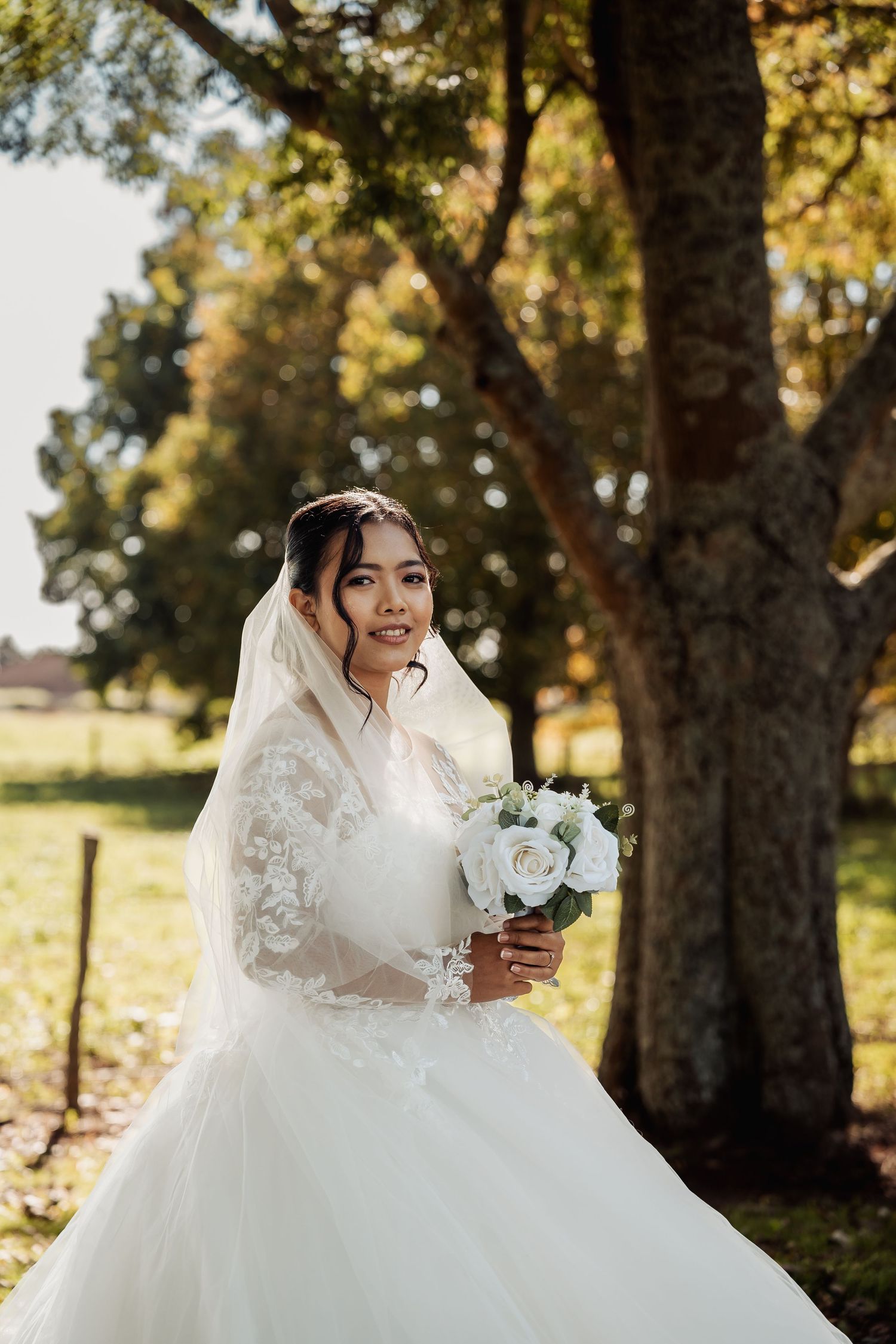 Radiant bride in lace wedding gown and veil holds white rose bouquet under golden autumn trees.