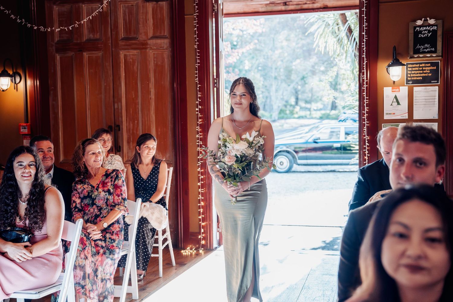 Wedding guests seated in a rustic wood-paneled venue watch as someone in a elegant gray gown enters with a bouquet.