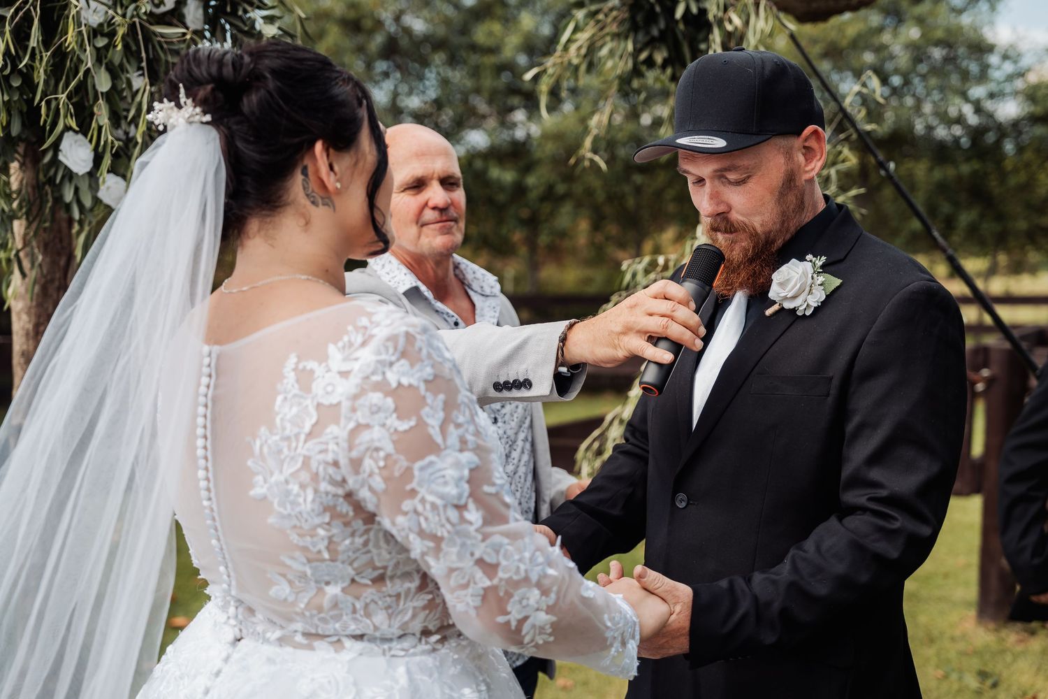 An outdoor wedding ceremony with a bride in lace long sleeve dress and groom in black suit exchanging rings.