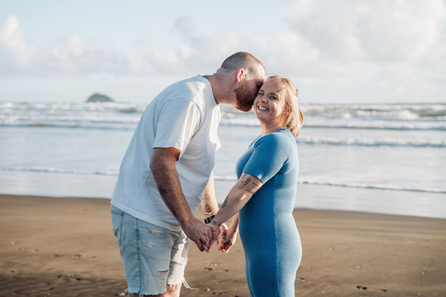 A couple embraces lovingly on a scenic beach at sunset with waves crashing in the background.