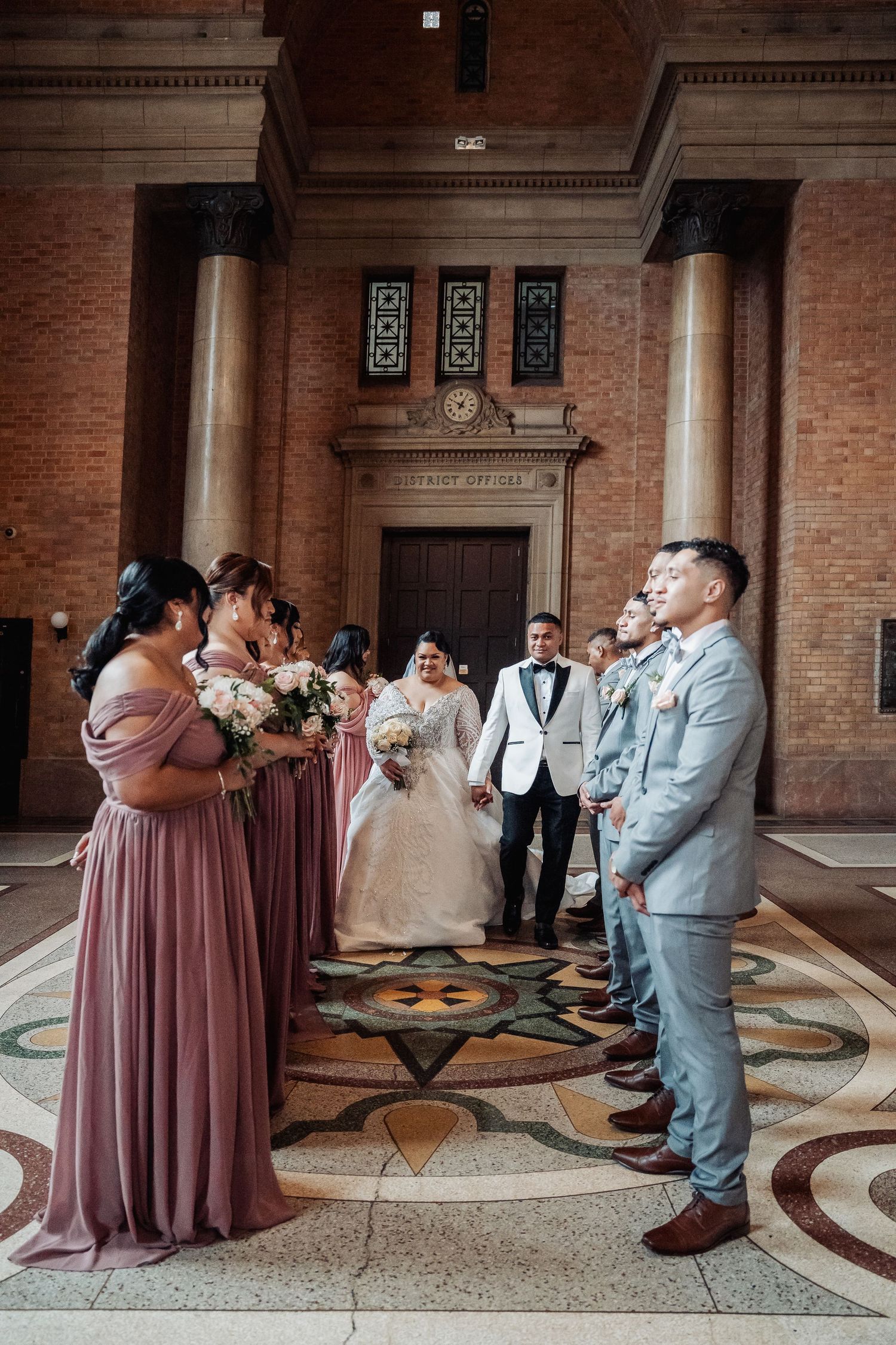Wedding party in dusty rose bridesmaids dresses and light gray suits gather on ornate tiled floor of historic brick building.
