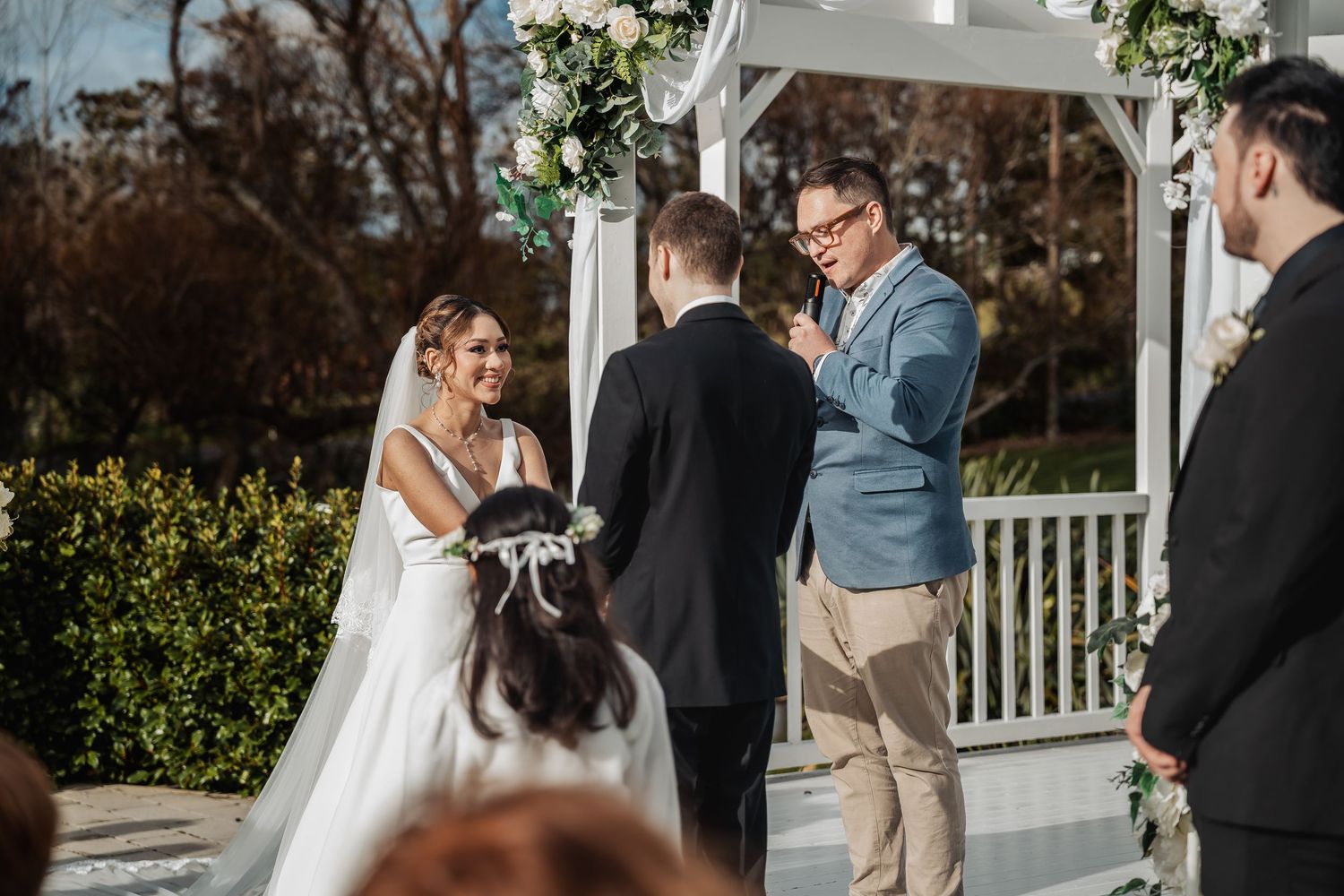 Wedding ceremony at an outdoor gazebo decorated with white flowers and greenery on a cloudy day.