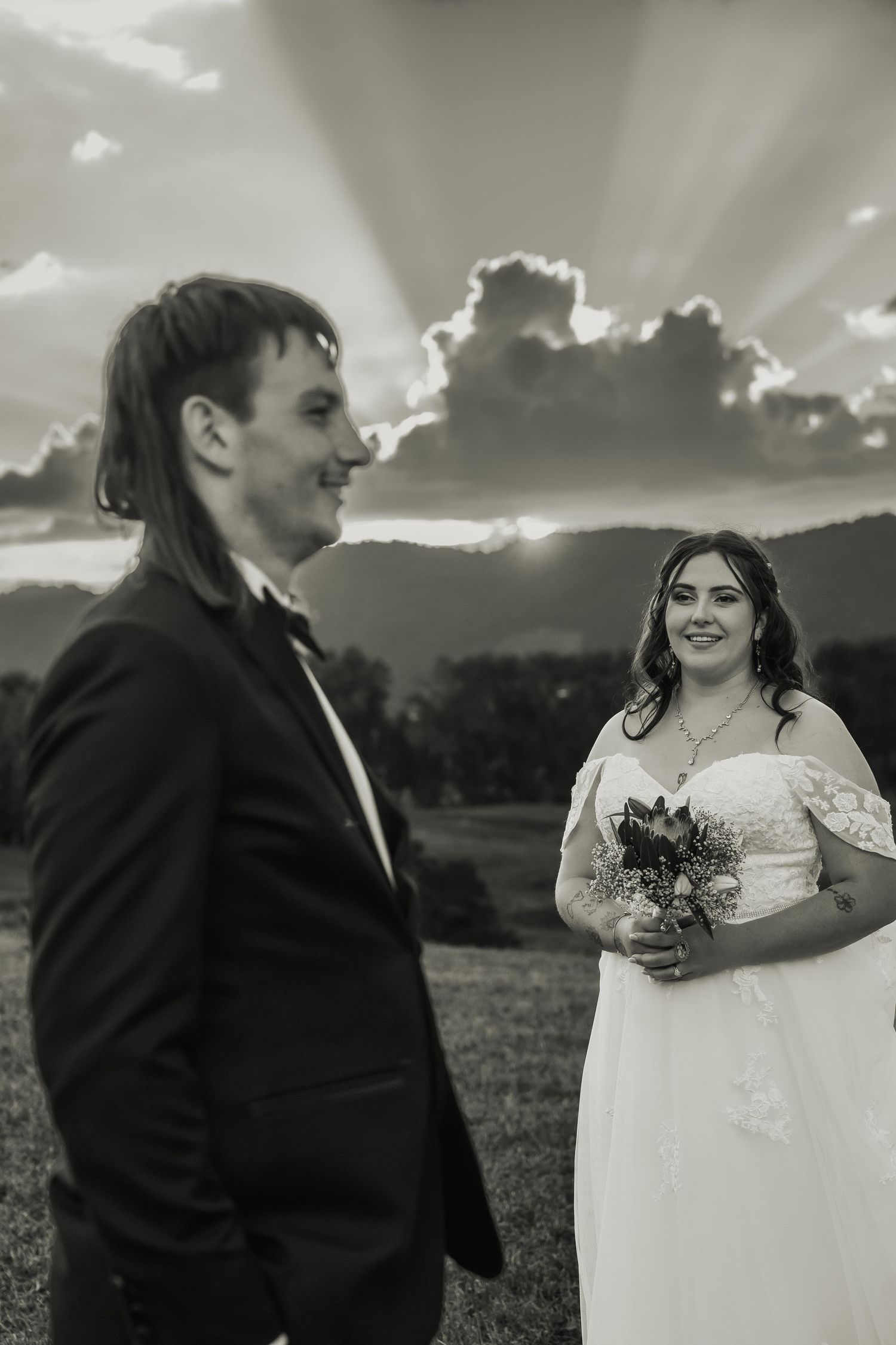 Black and white photo of bride holding bouquet smiling at groom with dramatic sunset rays behind clouds.