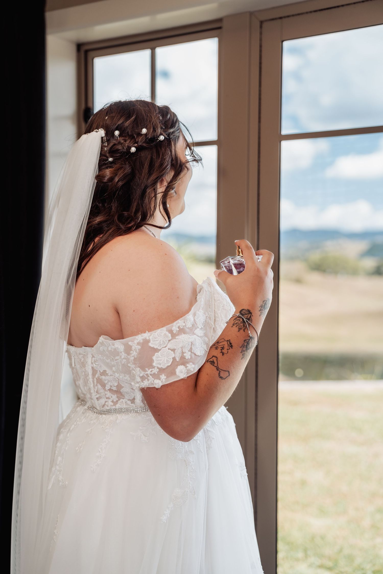 Bride in off-shoulder lace gown holding perfume bottle by window on wedding day.