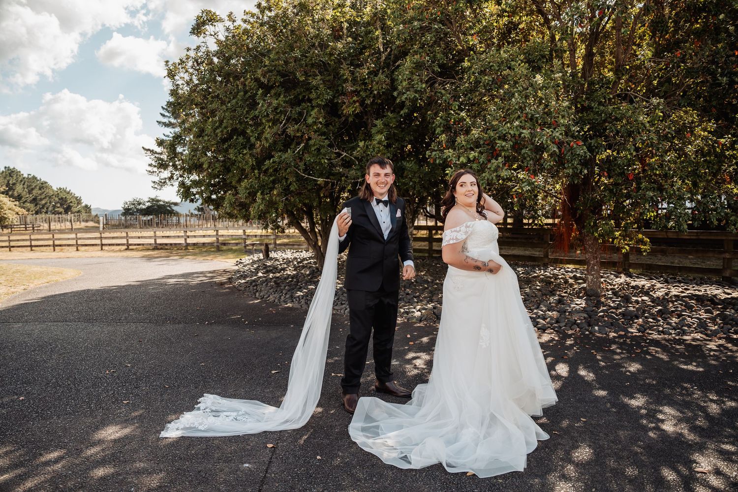 Bride and groom stand together on sunny driveway surrounded by trees at their outdoor wedding venue.