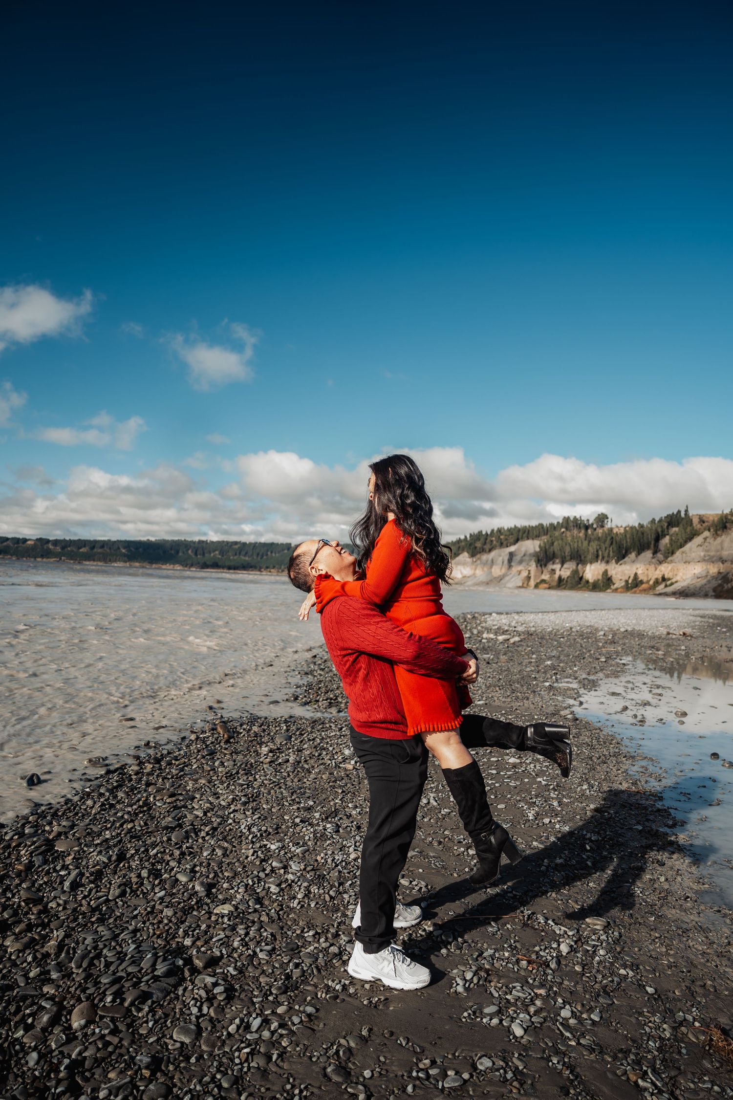 Man lifts woman in red dress on a sunny rocky beach with blue sky and cliffs in background.