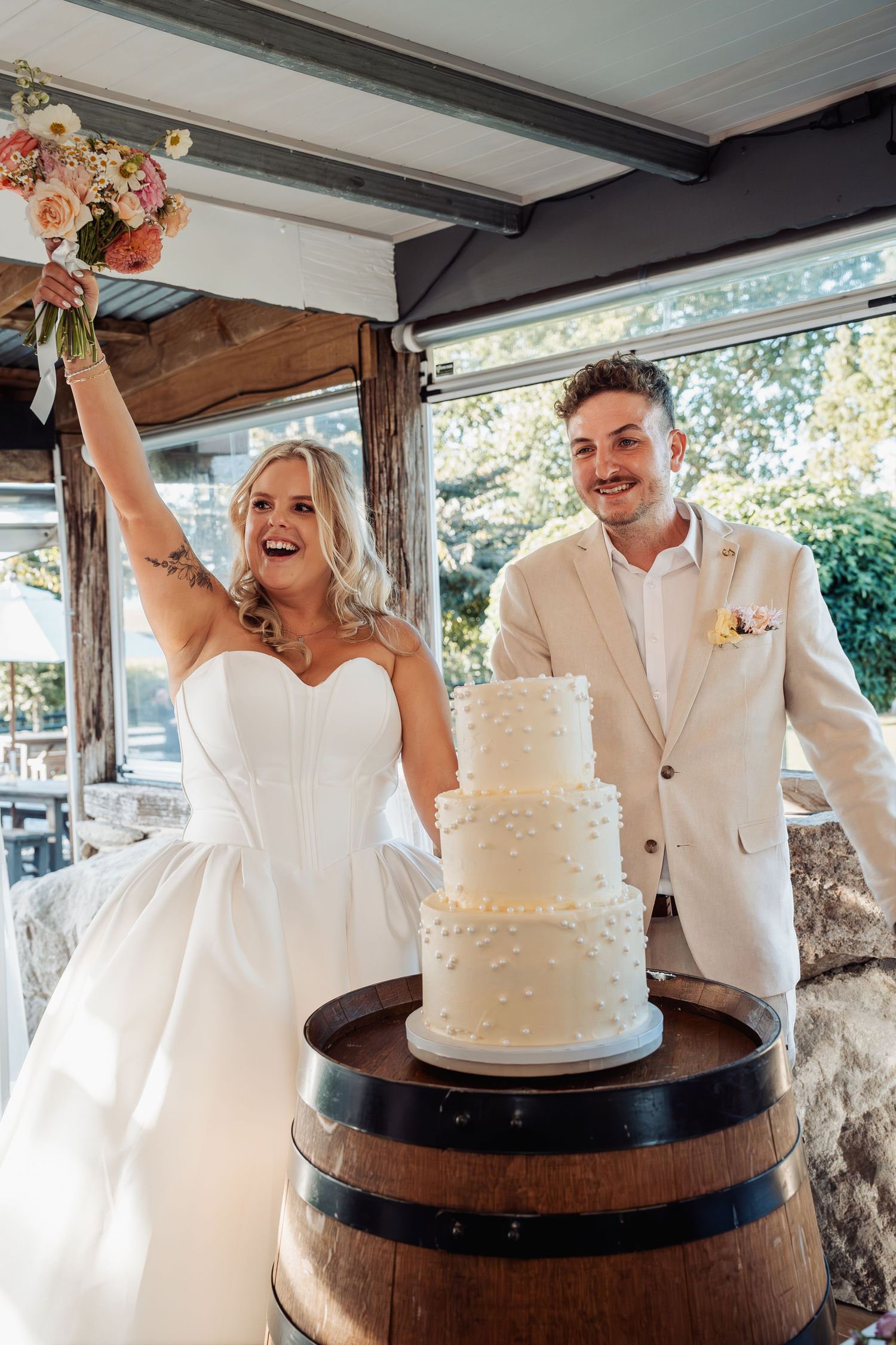 Joyful bride raises bouquet beside groom in front of three-tier white wedding cake on barrel.
