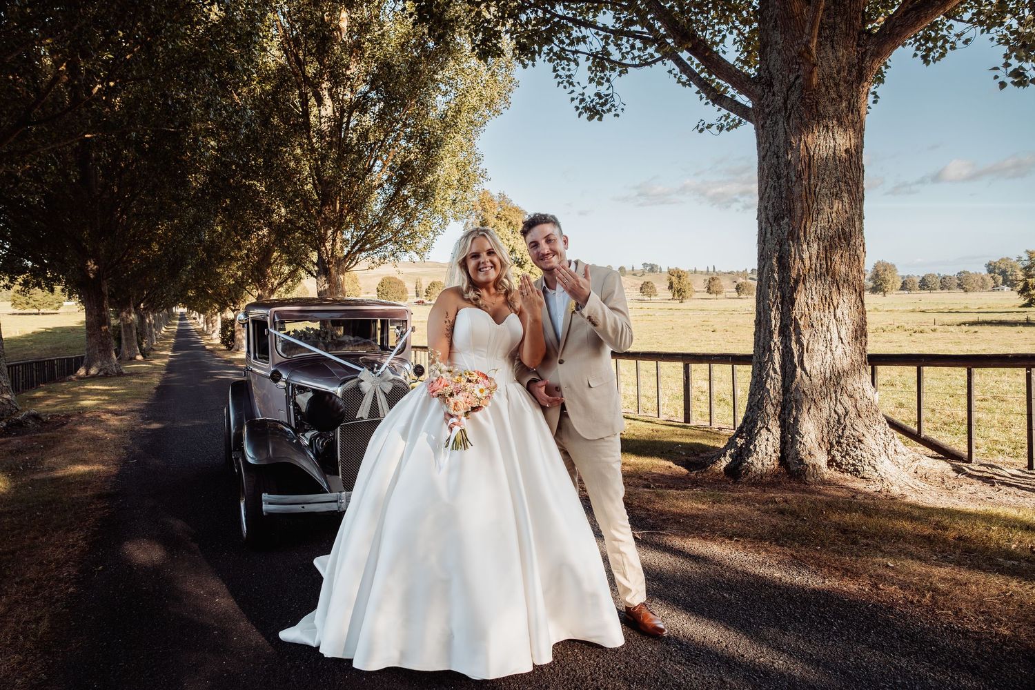Bride and groom posing beside a classic vintage car along a tree-lined driveway at sunset.