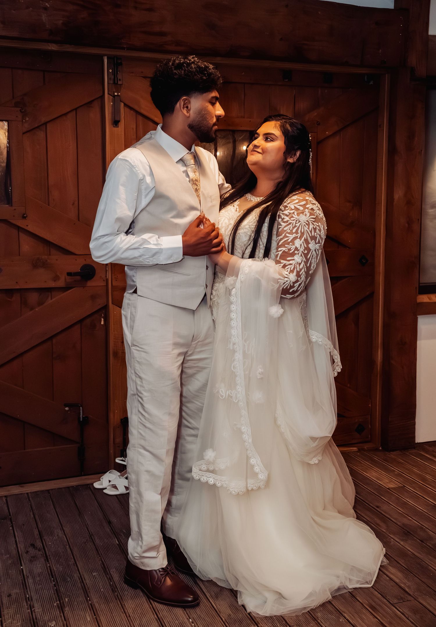 Newlyweds share intimate moment inside rustic barn venue, bride in lace gown holding grooms hands.