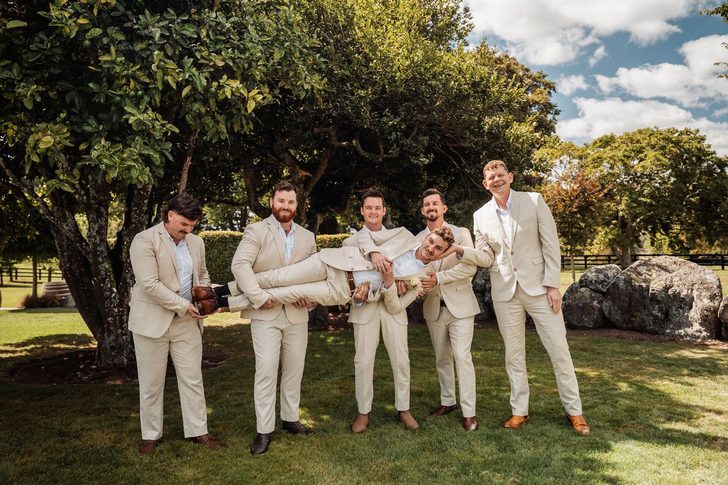 Groomsmen in beige suits playfully lifting groom in sunny garden with trees in background.