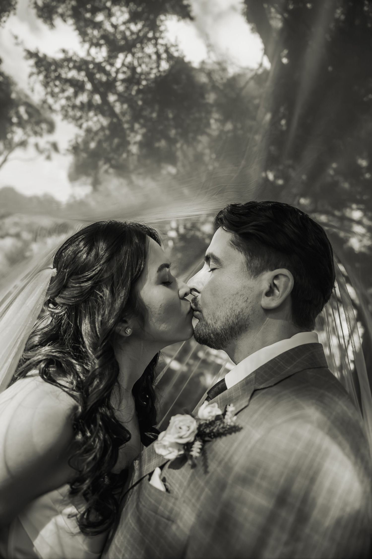 Black and white romantic portrait of bride and groom kissing beneath sheer wedding veil.