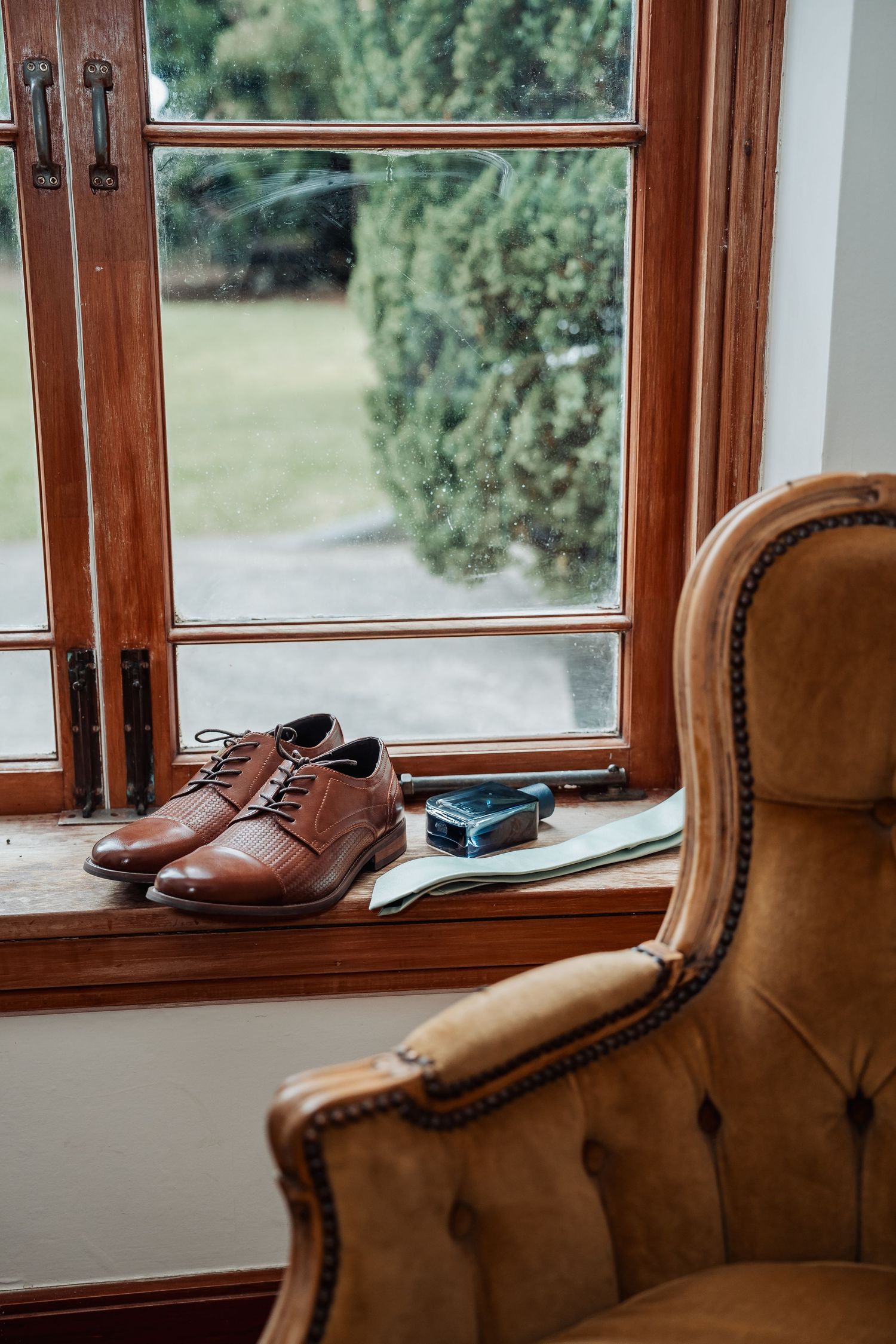 Brown leather dress shoes and a cologne bottle rest on a wooden windowsill beside a vintage armchair.