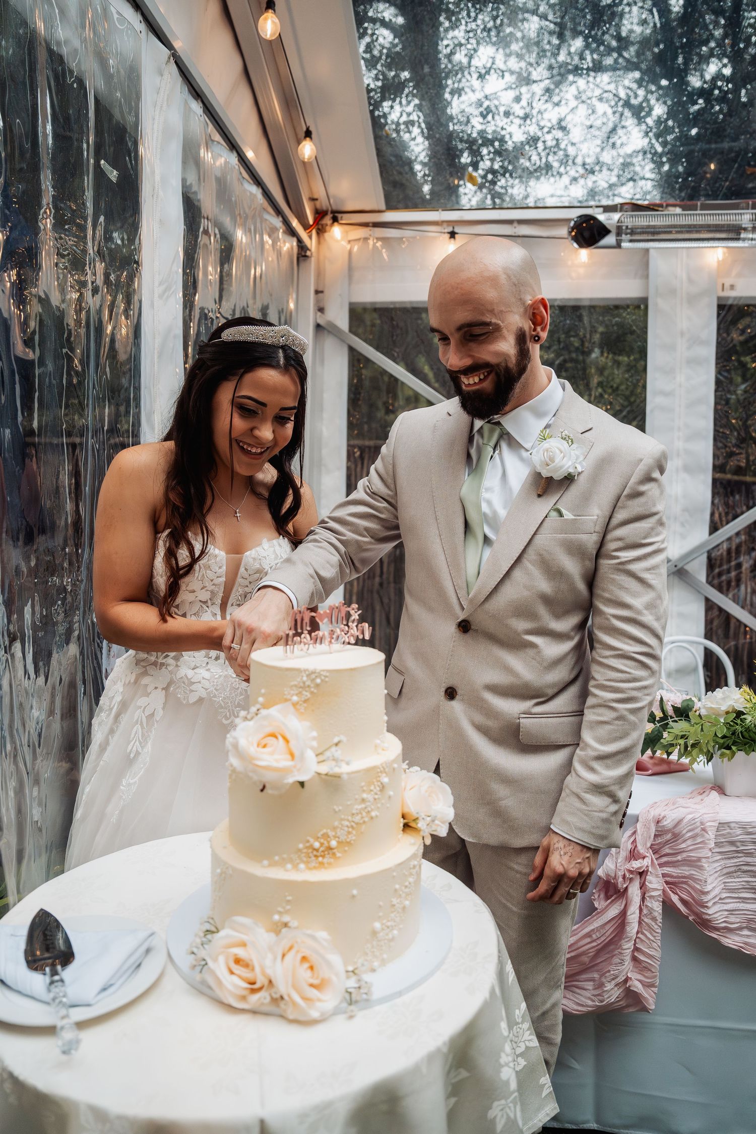 Smiling bride and groom cut three-tier white wedding cake decorated with roses together.