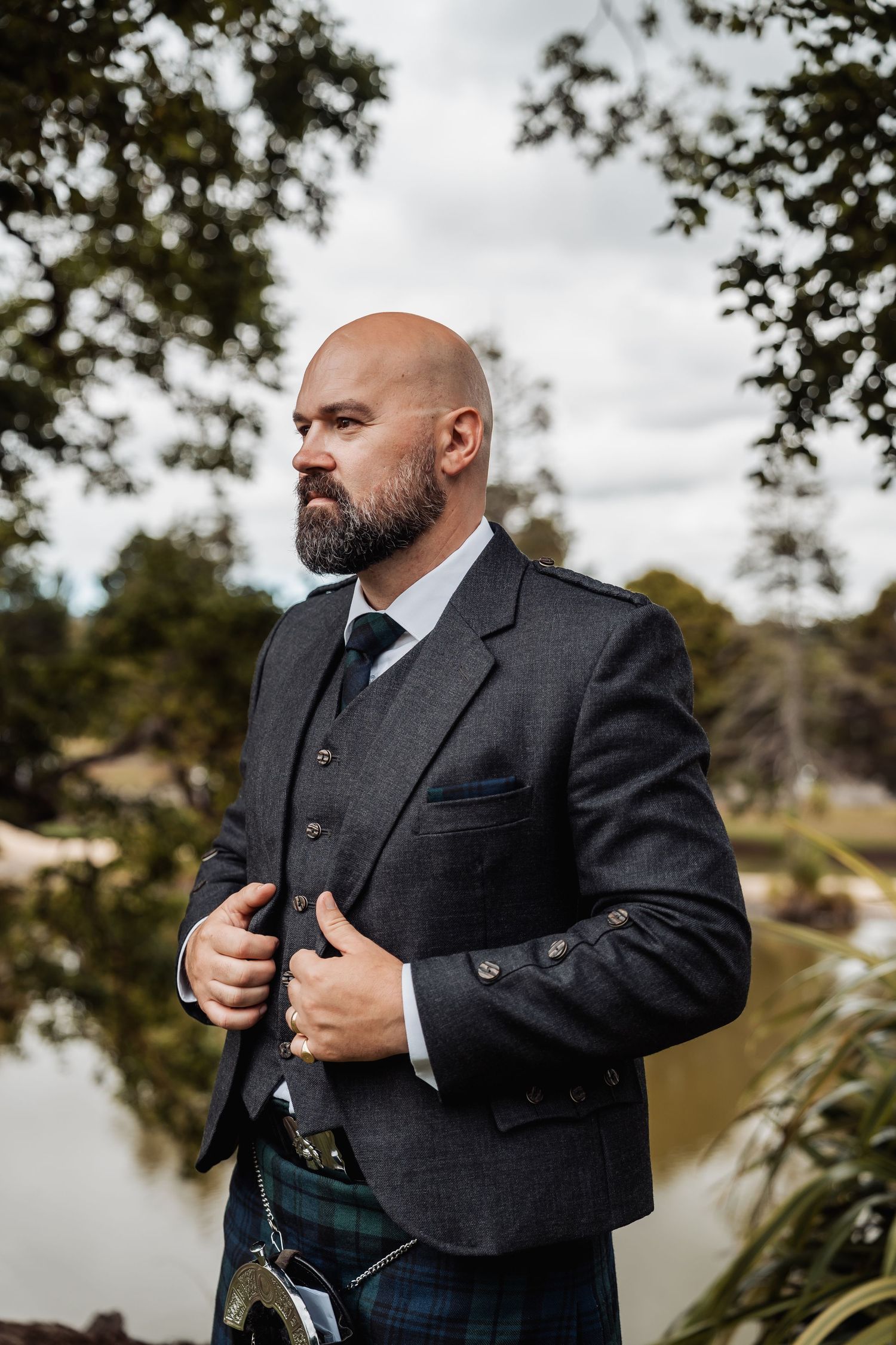 A bald bearded groom in a Scottish kilt and grey jacket poses confidently near a pond outdoors.