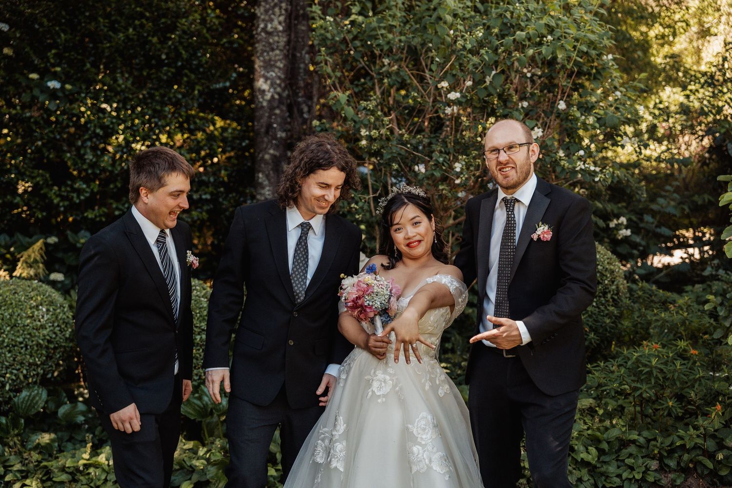 Bride in white lace gown holds bouquet with three suited groomsmen in lush garden setting.