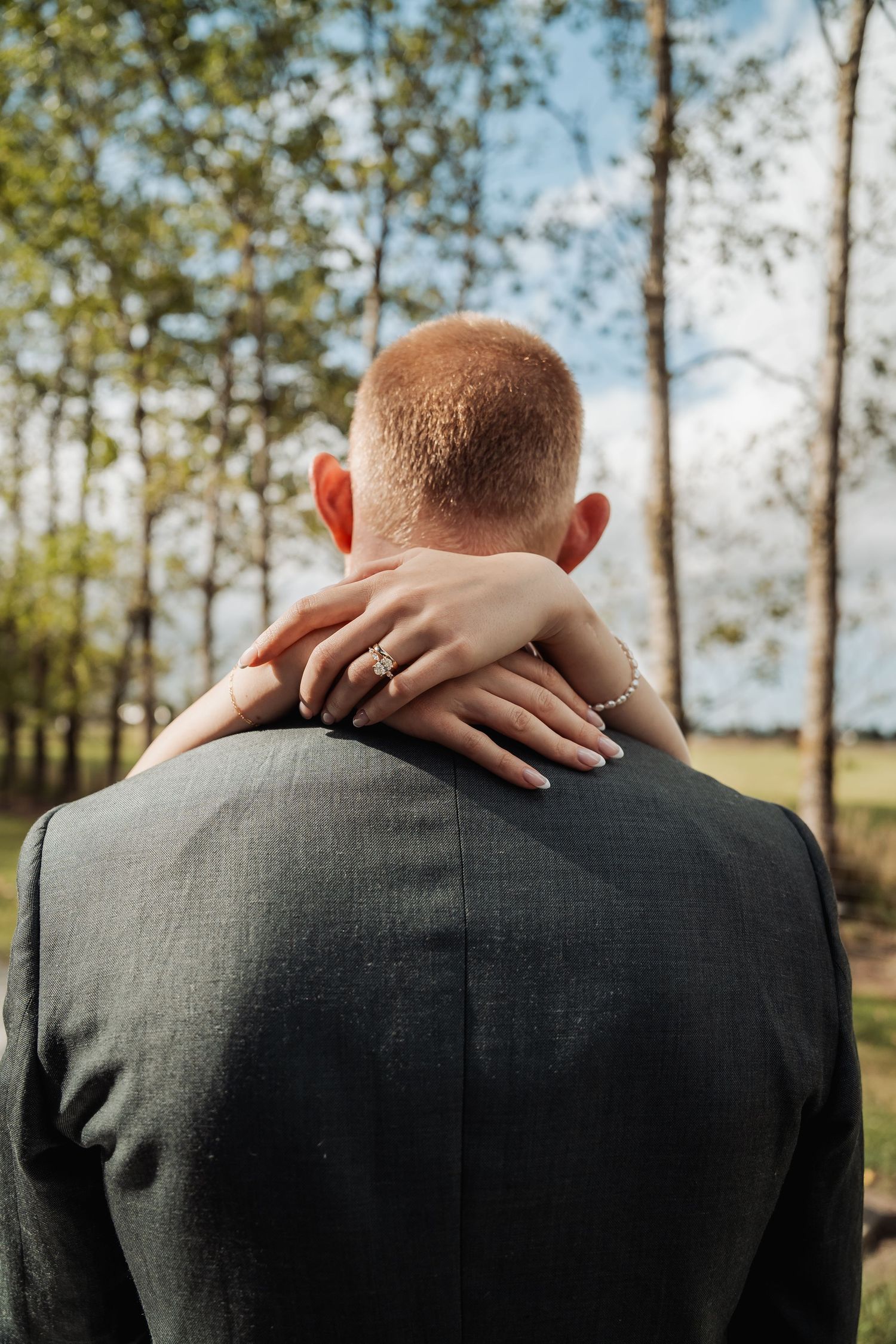Close-up of bride's hand with engagement ring resting on groom's shoulder from behind in a garden setting.