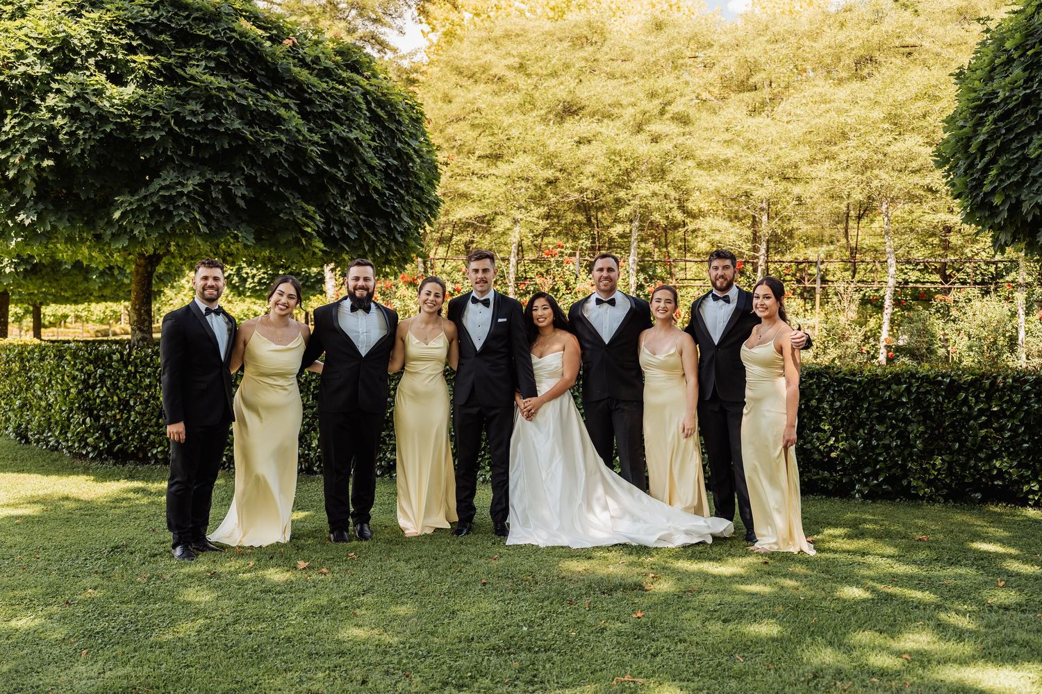 Wedding party in formal attire poses outdoors on green lawn with lush trees, bridesmaids in gold gowns and groomsmen in tuxedos.