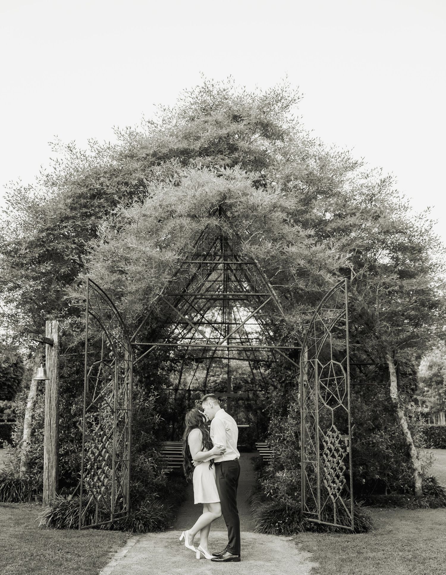 Couple kissing under ornate metal garden arch in black and white wedding photo.